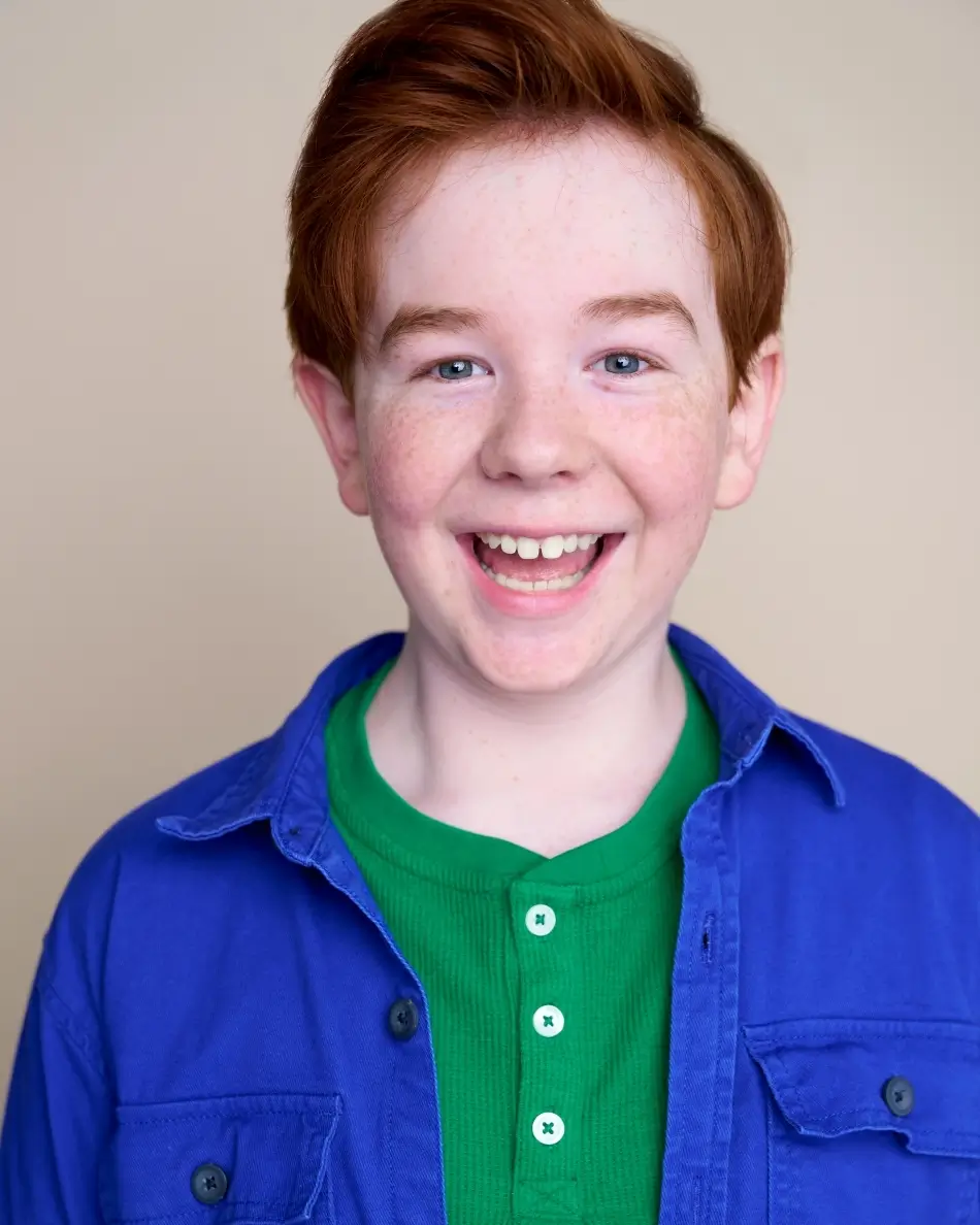 Atlanta kids actor headshot — young boy with red hair and freckles, bright smile in green shirt and blue button-up against neutral background, energetic fun commercial kids look.