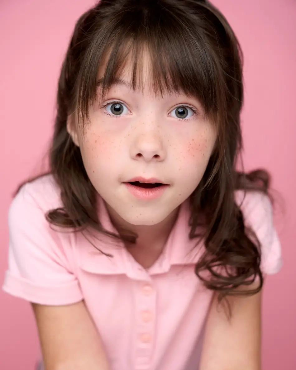 Atlanta kids actor headshot — young girl with brown hair and bangs, wide-eyed expressive look in pink polo shirt against pink background, theatrical kids character headshot.