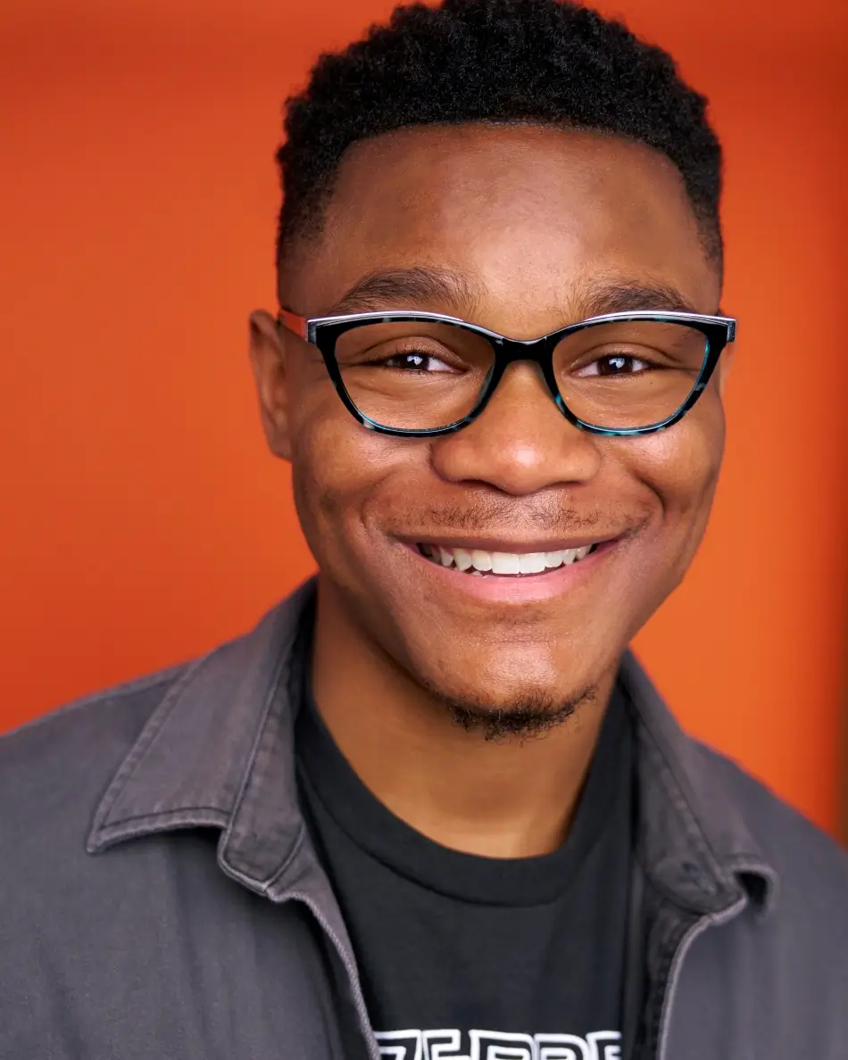 Atlanta teen/young adult actor headshot — male actor with glasses and warm smile in casual jacket and t-shirt against orange background, confident approachable commercial teen look.