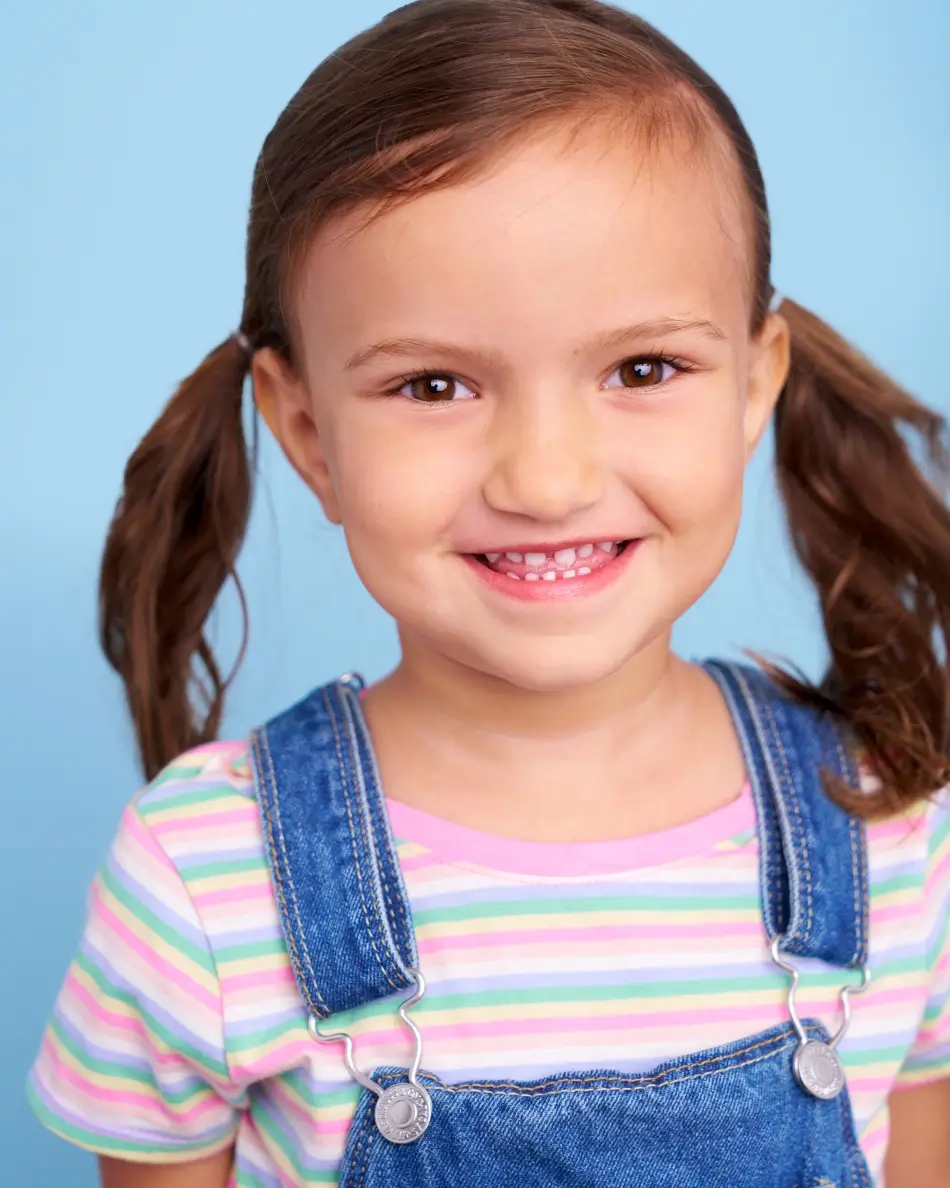 Atlanta kids actor headshot — young girl with brown hair in pigtails, big smile in striped shirt and denim overalls against light blue background, playful cheerful kids commercial look.