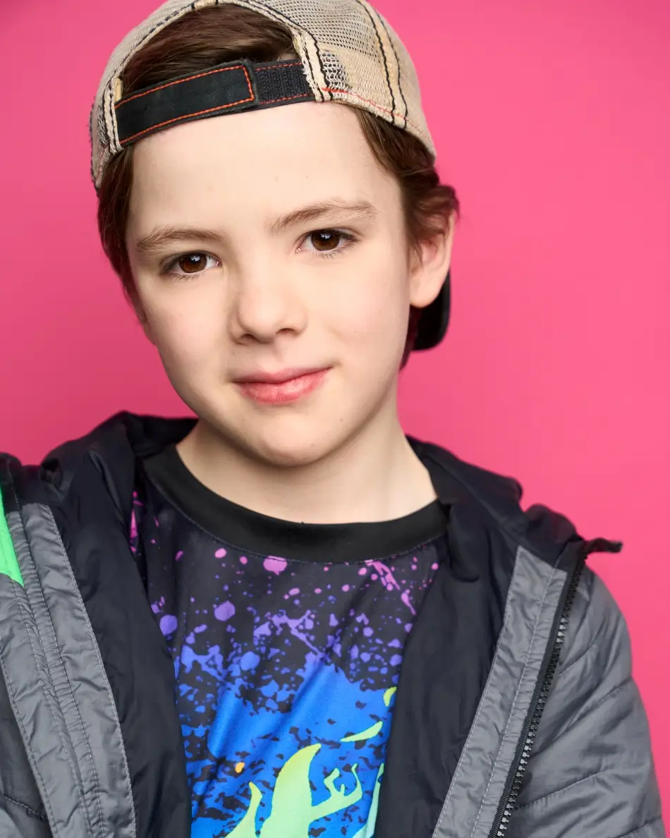 Atlanta kids actor headshot — young boy with baseball cap, confident expression in graphic t-shirt and jacket against pink background, theatrical kids character look.