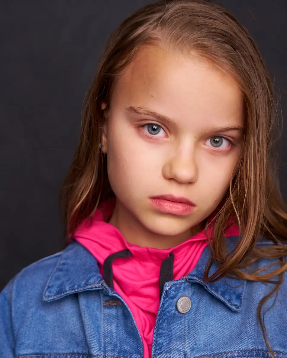 Atlanta kids actor headshot — young girl with long hair, intense serious expression in denim jacket and pink hoodie against dark background, theatrical drama-ready kids look.