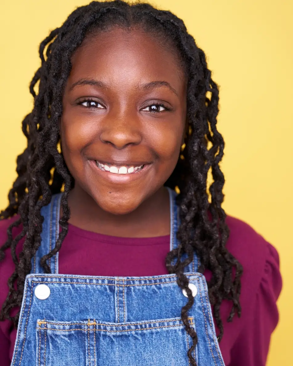 Atlanta kids actor headshot — young girl with long braids and big smile in burgundy top and denim overalls against yellow background, bright approachable kids commercial look.