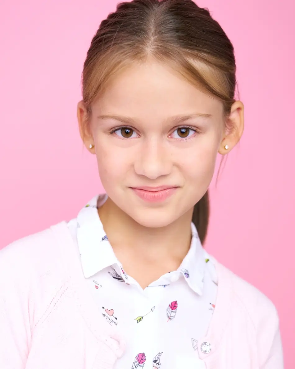 Atlanta kids actor headshot — young girl with light brown hair in ponytail, subtle smile in pastel pink sweater and floral collared shirt against pink background, confident soft commercial look.
