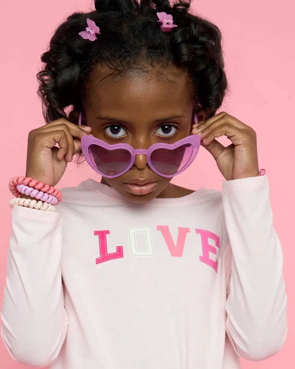 Atlanta kids actor headshot — young girl with styled hair and pink accessories, bold expression peeking over heart-shaped sunglasses in LOVE shirt against pink background, playful kids commercial character look.
