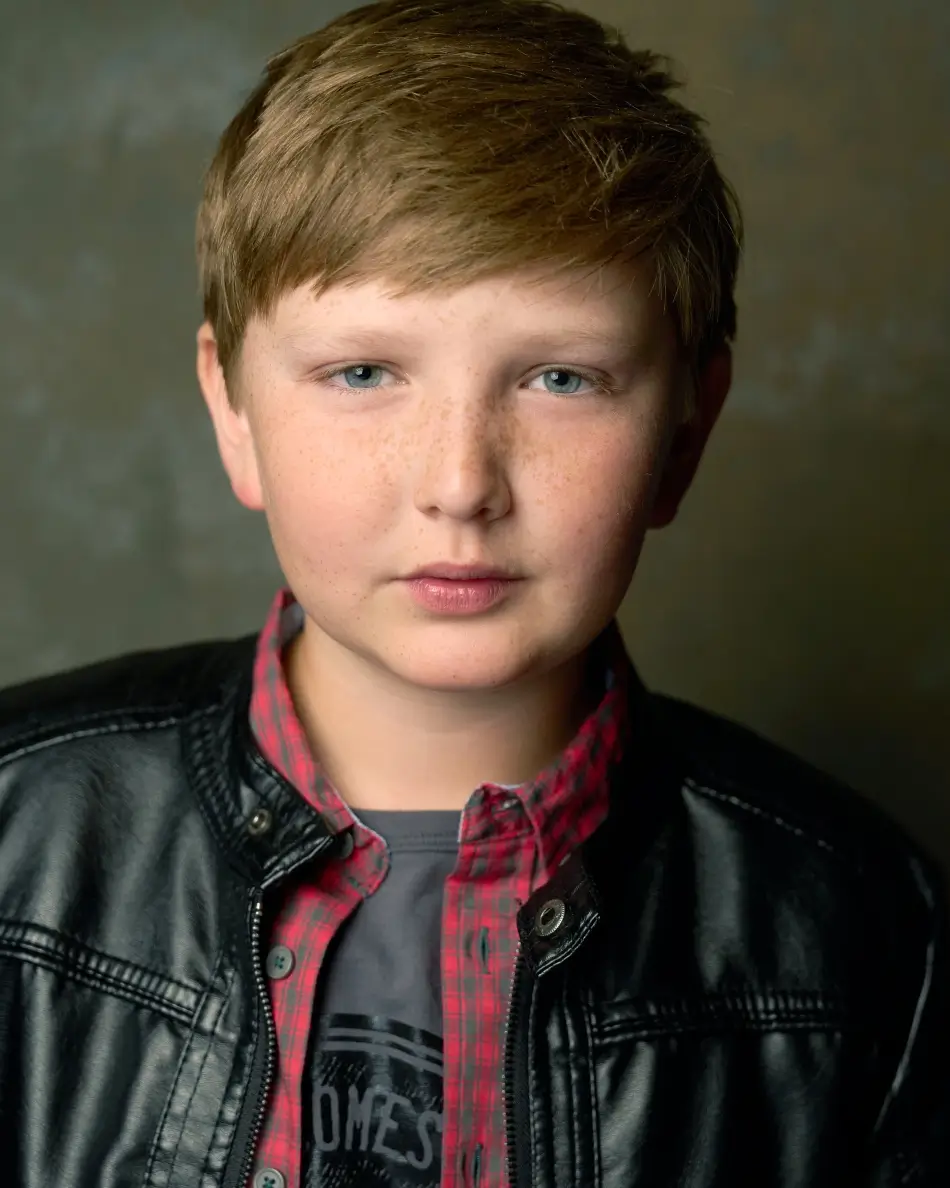 Atlanta kids actor headshot — young boy with light brown hair and freckles, serious expression in leather jacket, plaid shirt, and graphic tee against neutral backdrop, grounded theatrical drama kids look.