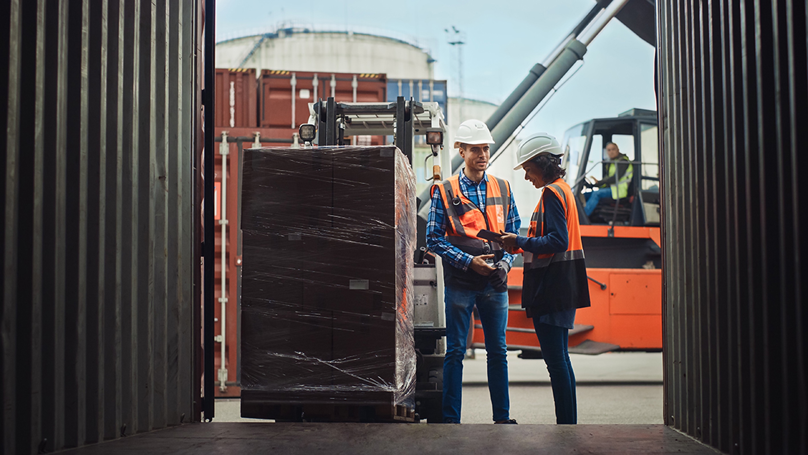 Warehouse workers in safety vests and hard hats coordinate a pallet delivery with a forklift at a loading dock, reviewing shipment details on a handheld device.