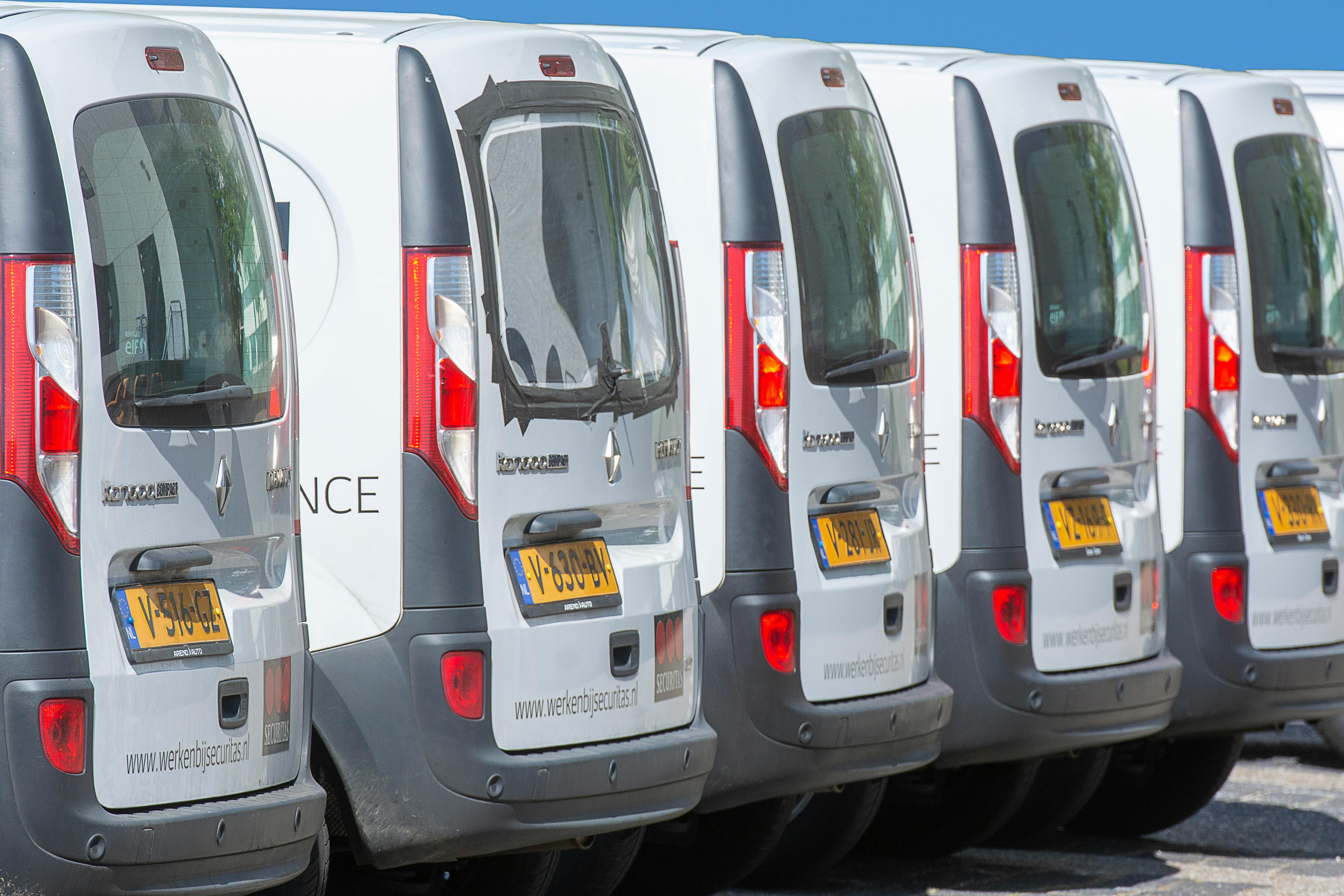 A row of white delivery vans parked side by side in a parking lot, viewed from the rear.
