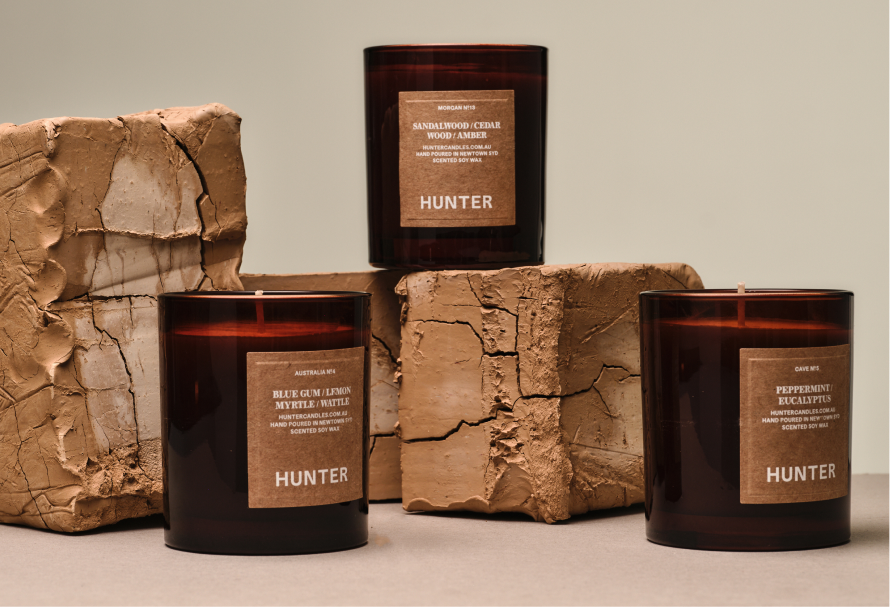 Three amber glass scented candles with brown labels reading "HUNTER" are displayed on cracked clay blocks against a warm neutral background. One candle is elevated at the back, while two sit in the foreground. The labels feature different fragrance blends, including Sandalwood/Cedar Wood/Amber, Blue Gum/Lemon Myrtle/Wattle, and Peppermint/Eucalyptus, creating an earthy, premium product presentation.