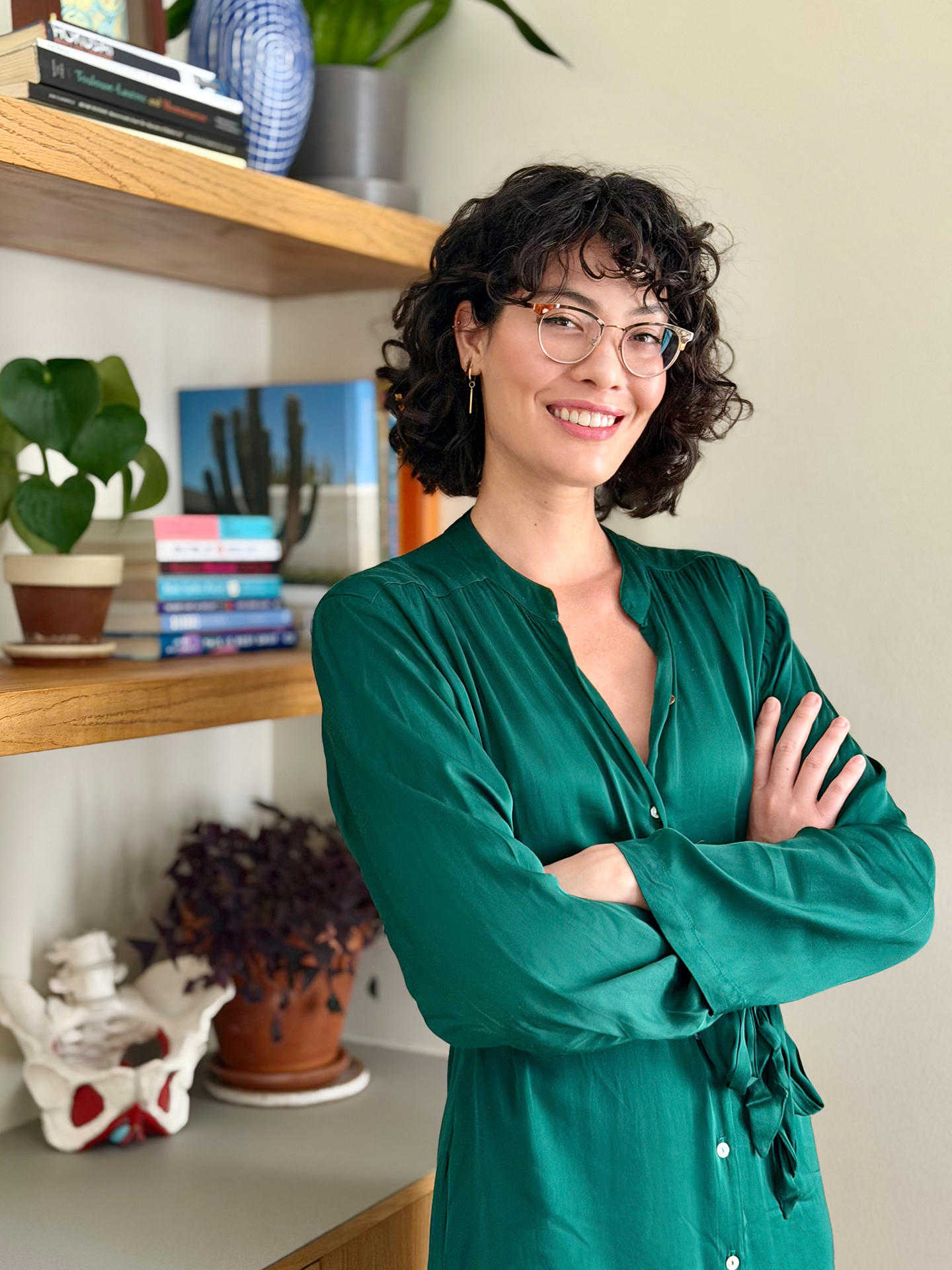 Headshot of Anna Kimura smiling with crossed arms in her office, featuring an anatomical pelvic model in the background.