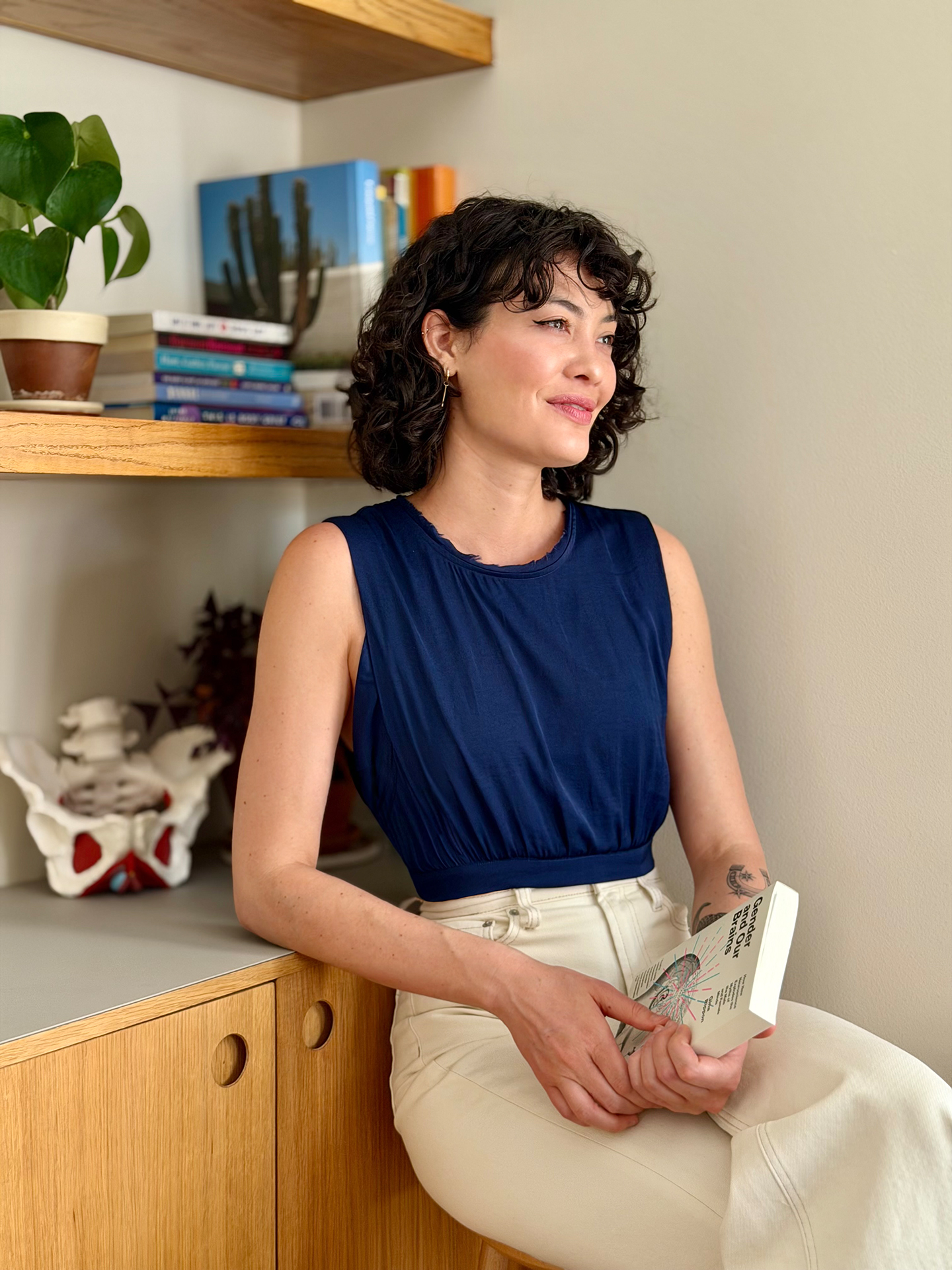 Anna Kimura sitting by a wooden clinic bookshelf, holding a book in her lap with stacked reference books and a plant on the shelves behind her.