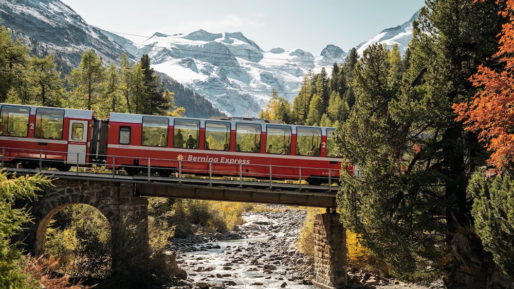 Red Bernina Express train crossing a stone bridge over a rocky river with snowy mountains in the background.
