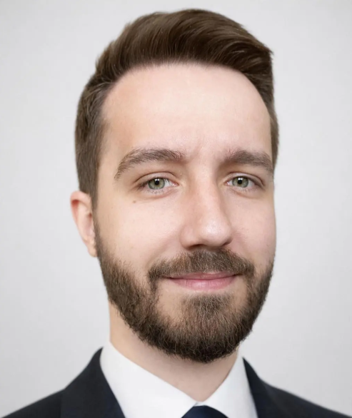 Mężczyzna z krótkimi brązowymi włosami i brodą, ubrany w czarny garnitur, białą koszulę i ciemny krawat, uśmiechający się na jasnym tle.