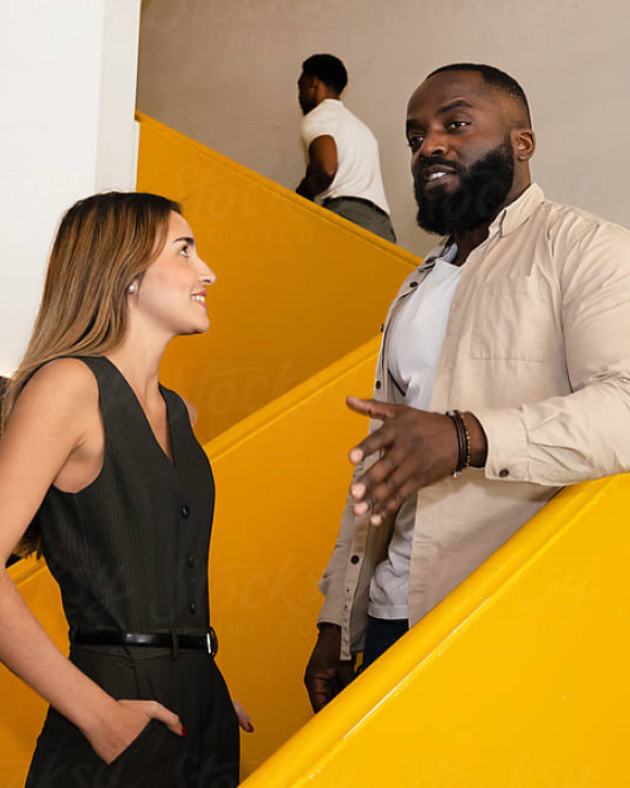 
A man and woman are talking on a bright yellow staircase. The man on the right, who has a beard and is wearing a tan jacket over a white shirt, gestures with his hands as he speaks. The woman on the left, who is wearing a black pinstripe vest and pants, looks at him and smiles. In the background, another man walks up the stairs.