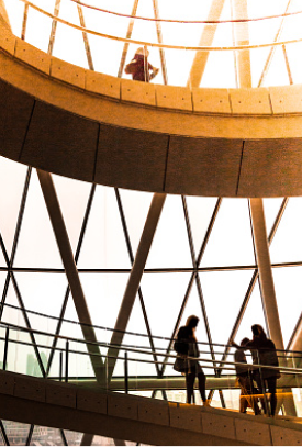 A blurry, high-contrast, low-angle shot of the inside of a tall, domed building. The image is taken from a low angle, looking up. The building has a curved, skeletal structure of metal or concrete beams that create large, diamond-shaped windows, and a spiraling walkway. Silhouetted figures of people are visible on the different levels of the walkway. The light from outside is bright, making the interior dark and the people look like silhouettes.