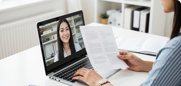 Person holding a document and participating in a video call on a laptop with a smiling woman on screen.