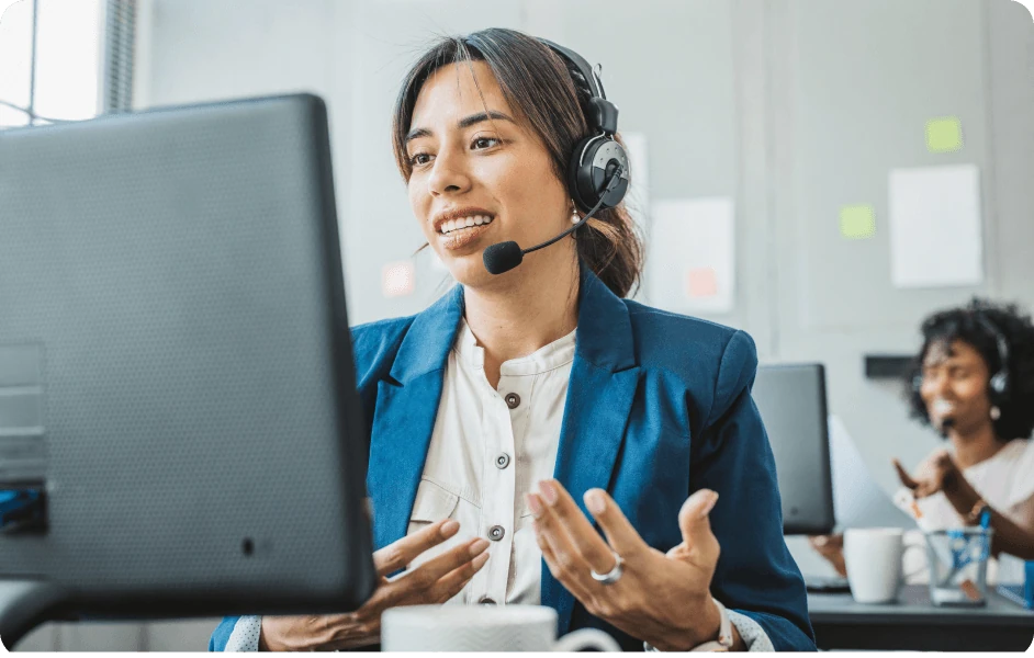 Professional woman wearing headset and blue blazer speaking in front of a computer during a video call in an office setting.