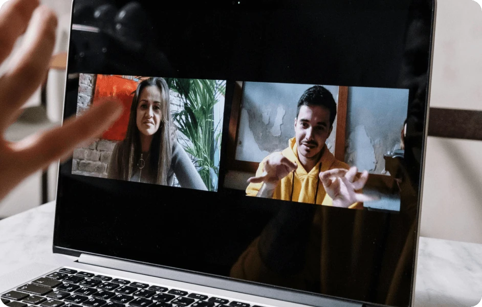 Two people on a laptop screen video call, a woman on the left and a man in a yellow hoodie on the right, with a hand waving in front of the laptop.