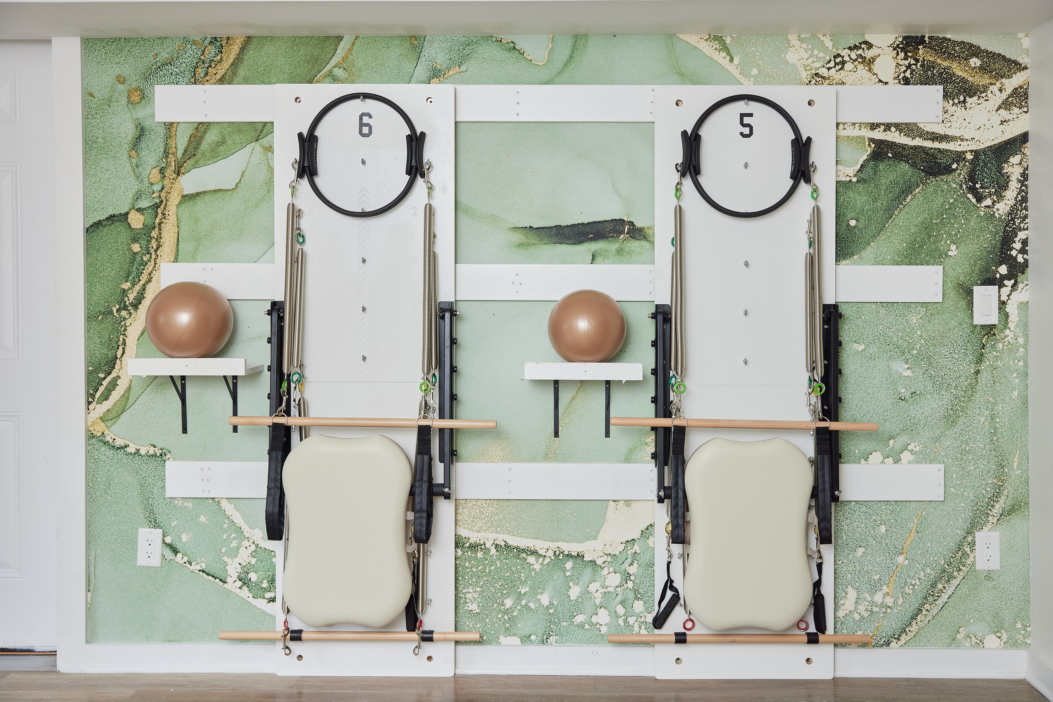 Two Pilates wall units with springs, wooden bars, and padded backrests mounted on a green and beige marbled wall, each holding a bronze exercise ball on a small shelf.