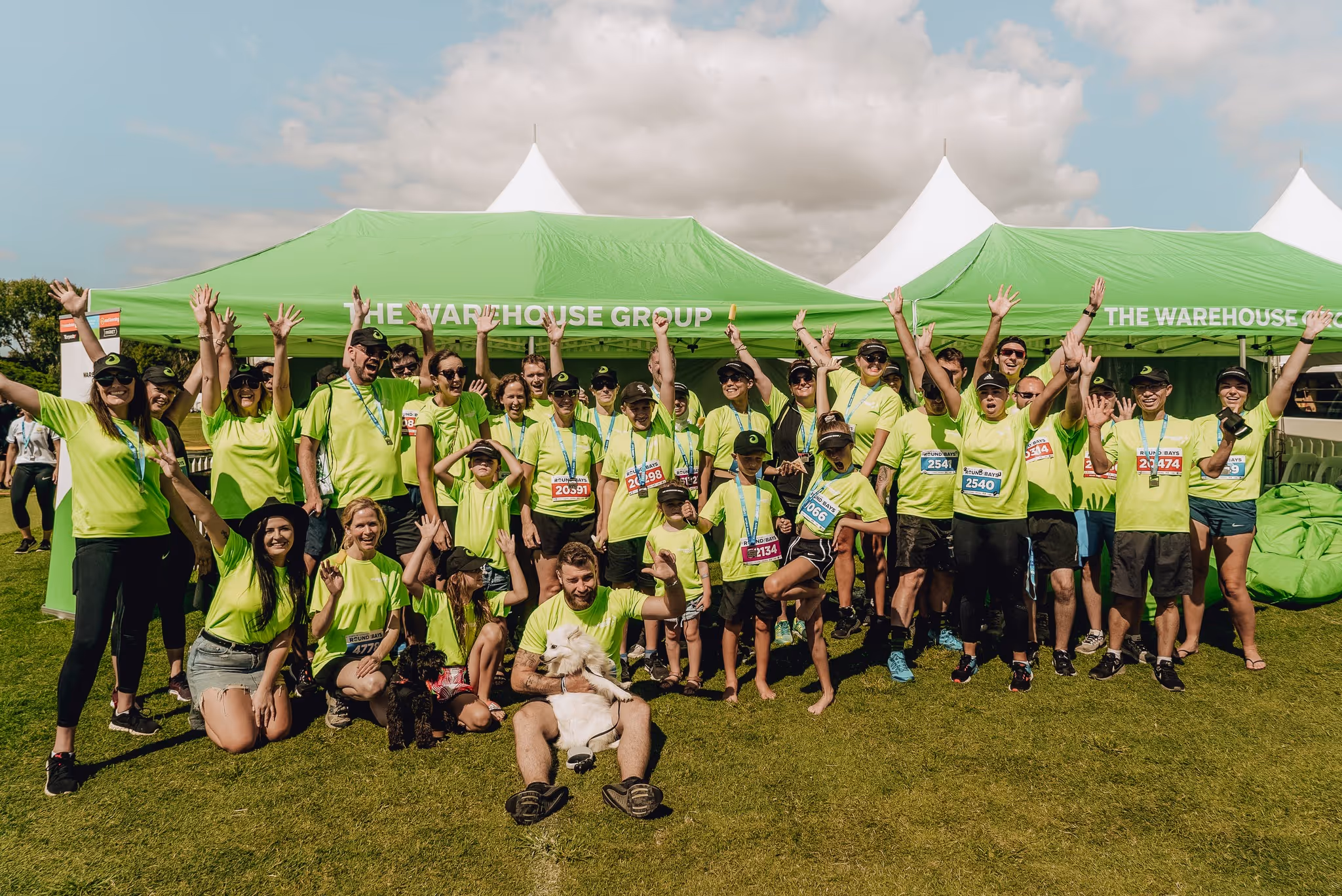 A group of people wearing matching bright green shirts pose enthusiastically with arms raised in front of large green tents on a sunny day. The group looks joyful and united, likely participating in an outdoor event or activity.