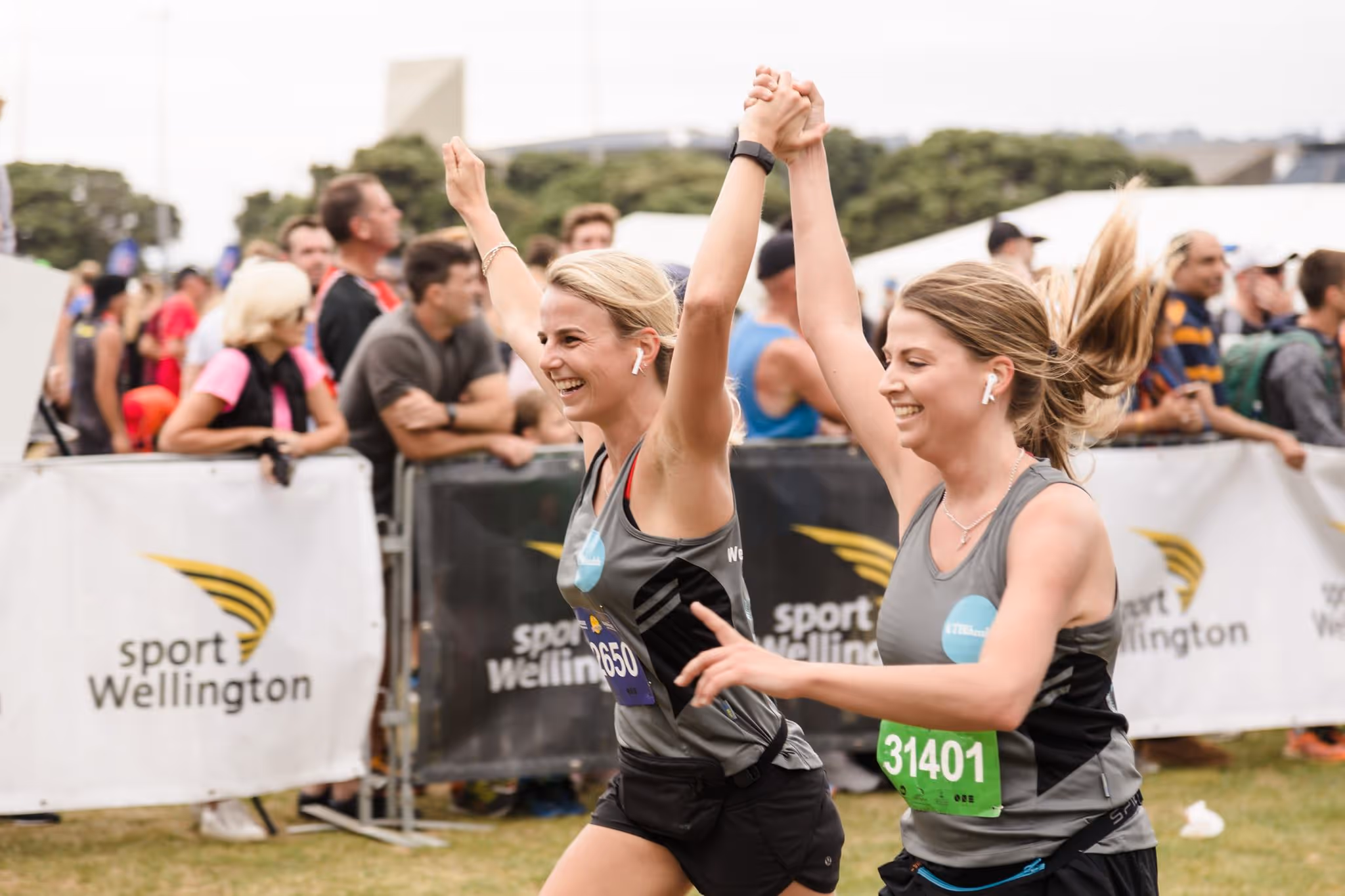 Two women in athletic gear cross the finish line of a race, holding hands and raising their arms in celebration. They are surrounded by cheering spectators and banners that say "Sport Wellington." The atmosphere is jubilant, and both women appear elated.