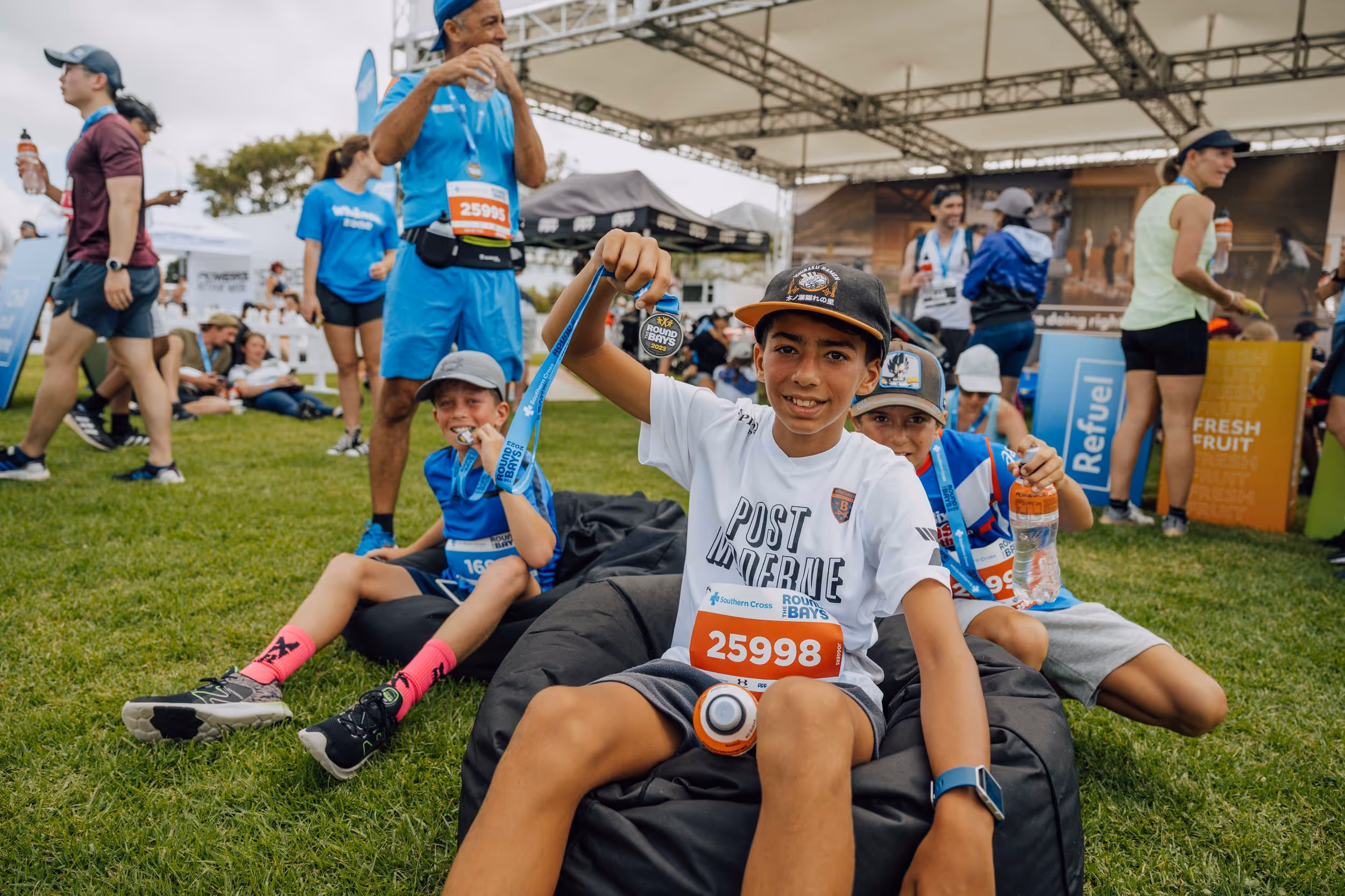 A group of people, including children and adults, are outdoors under a canopy. The central child, wearing a white t-shirt and cap, is smiling and raising their fist triumphantly while holding a medal. Others around are sitting or standing, enjoying the event.