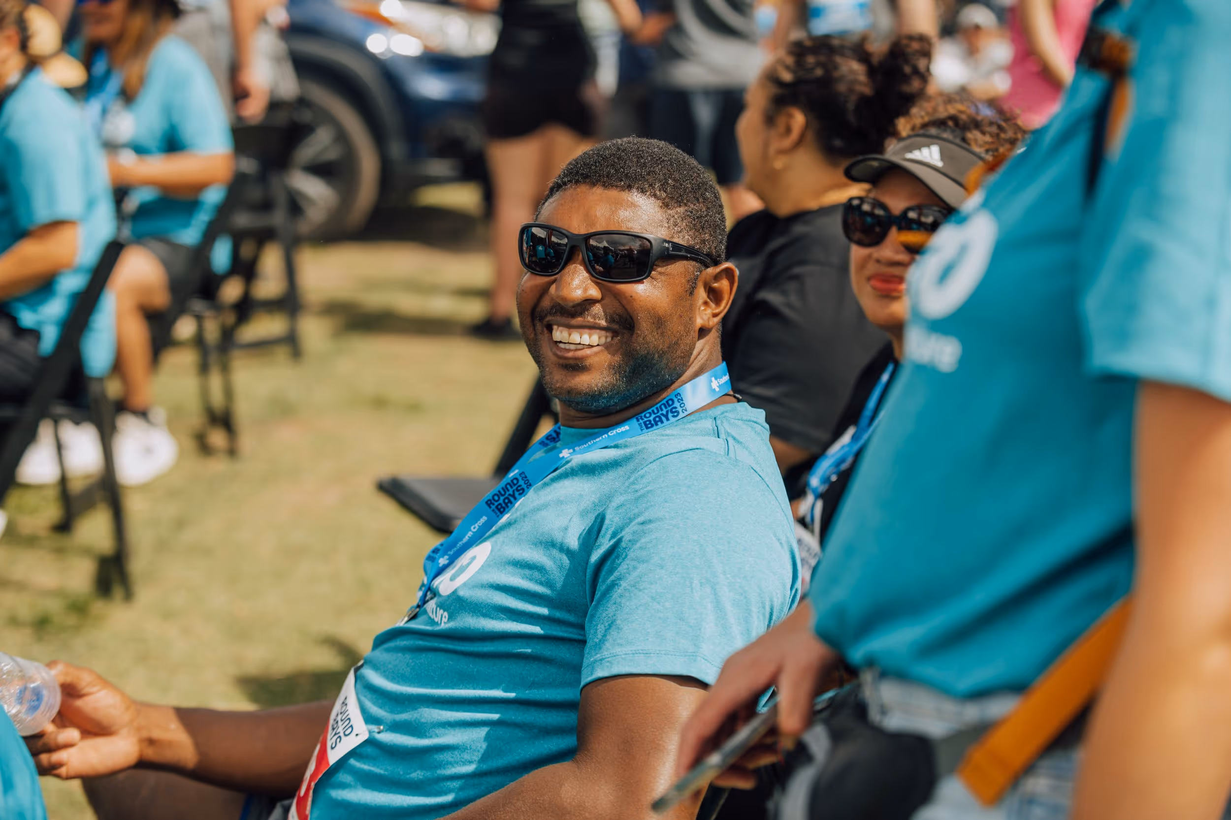A man wearing sunglasses and a teal shirt with a lanyard smiles at the camera while sitting outdoors among a group of people. Others around him are also wearing similar teal shirts, suggesting they are part of the same event or organization.