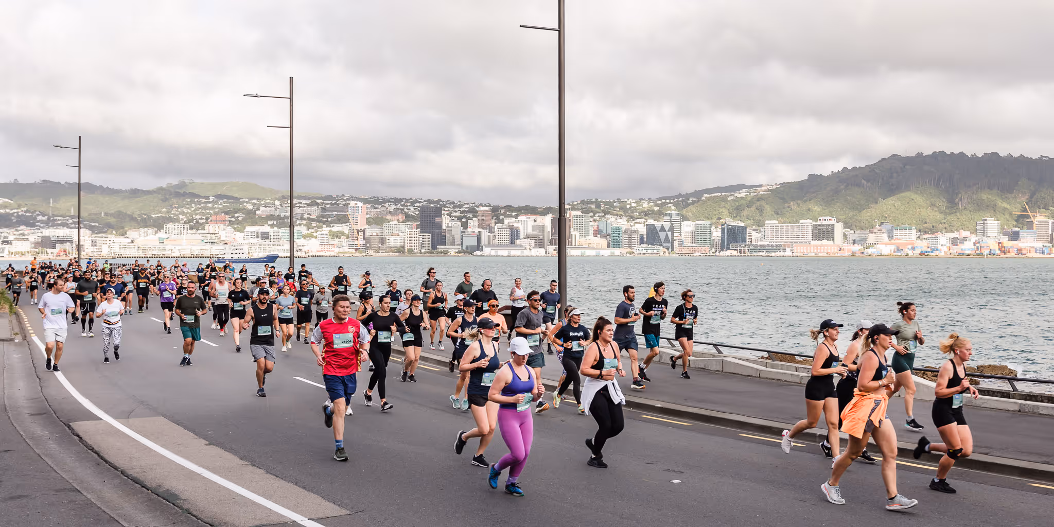 A large group of runners, including men and women, participates in a marathon along a seaside road. The sky is overcast, and there are hills and buildings visible in the distance across the water. The road has light posts, and some people watch from the sidelines.