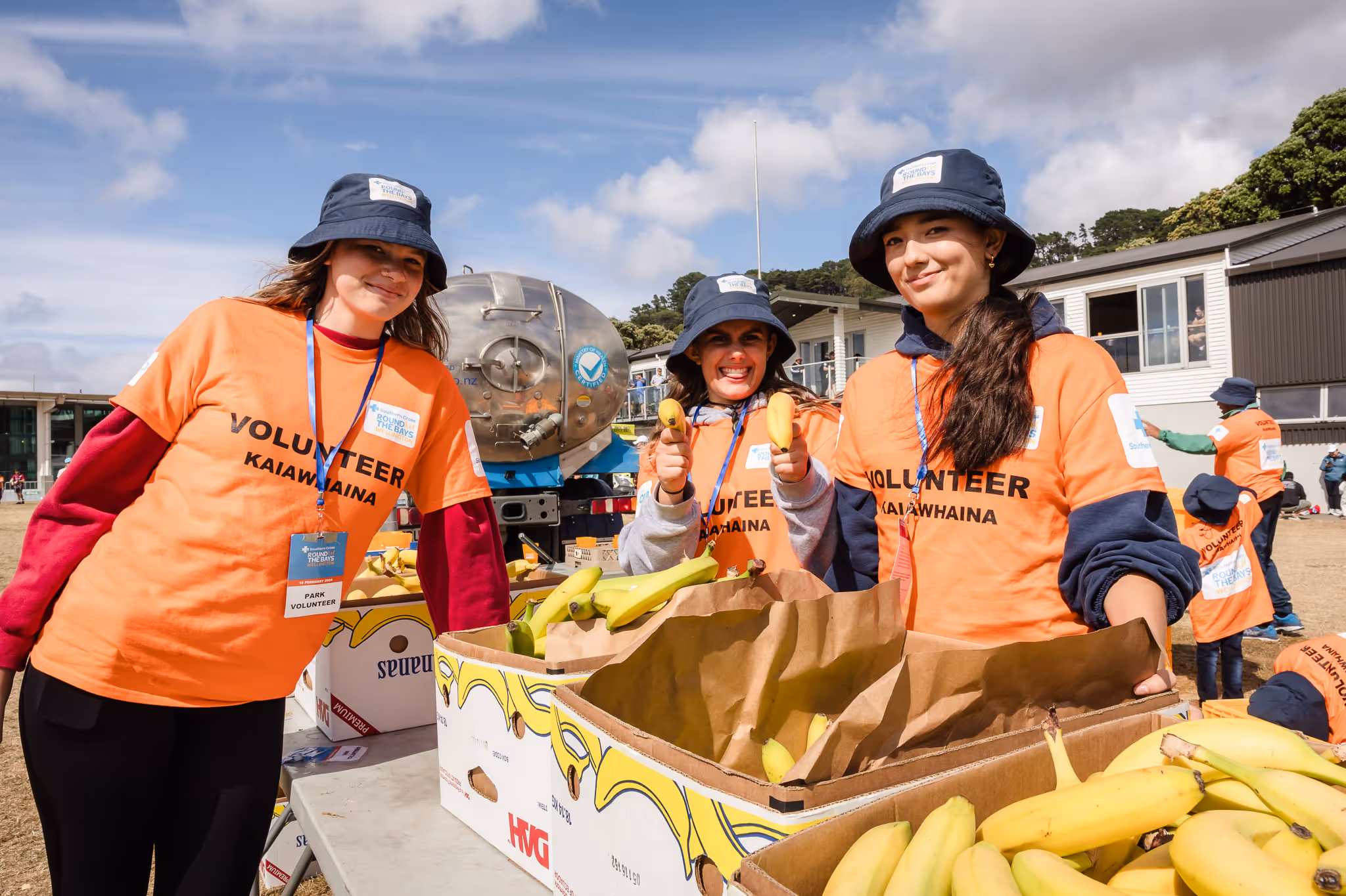 Three people wearing orange shirts and blue hats are smiling and distributing bananas at an outdoor event. They stand behind a table with a large box full of bananas. Other event participants can be seen in the background.