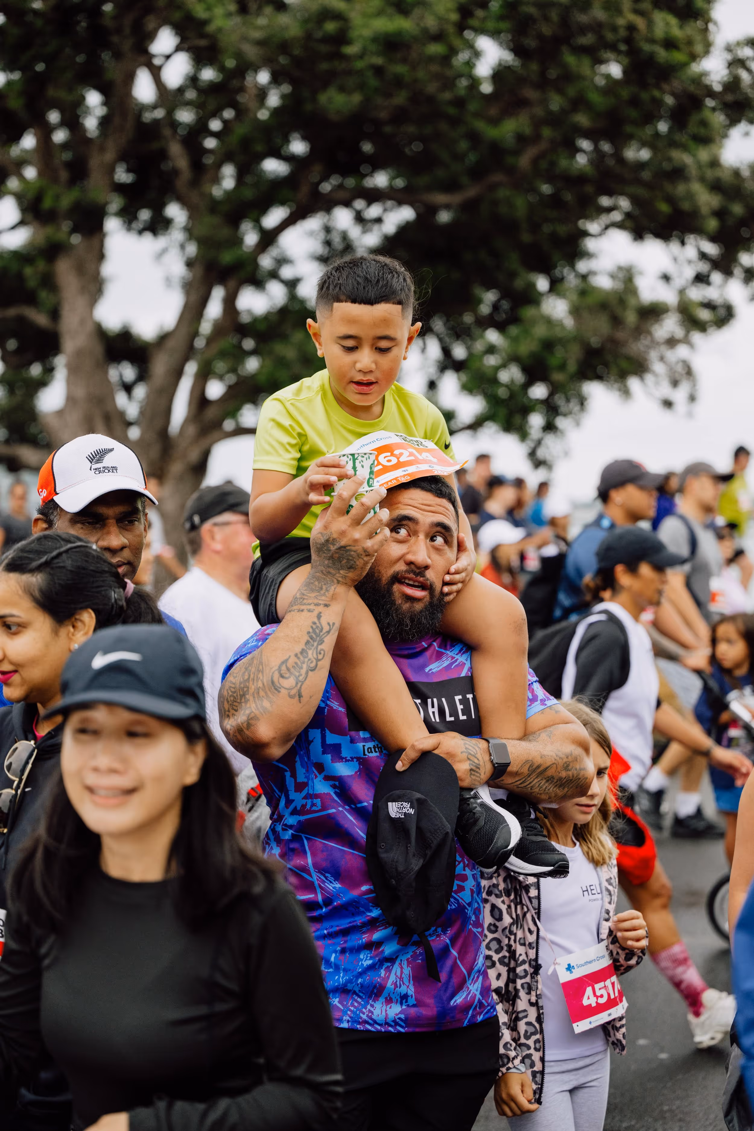 A man carries a child on his shoulders among a crowd of people walking outdoors. The child wears a yellow shirt, and the man is in a purple tie-dye shirt and has visible tattoos. The background shows trees and more participants in what appears to be a community event.