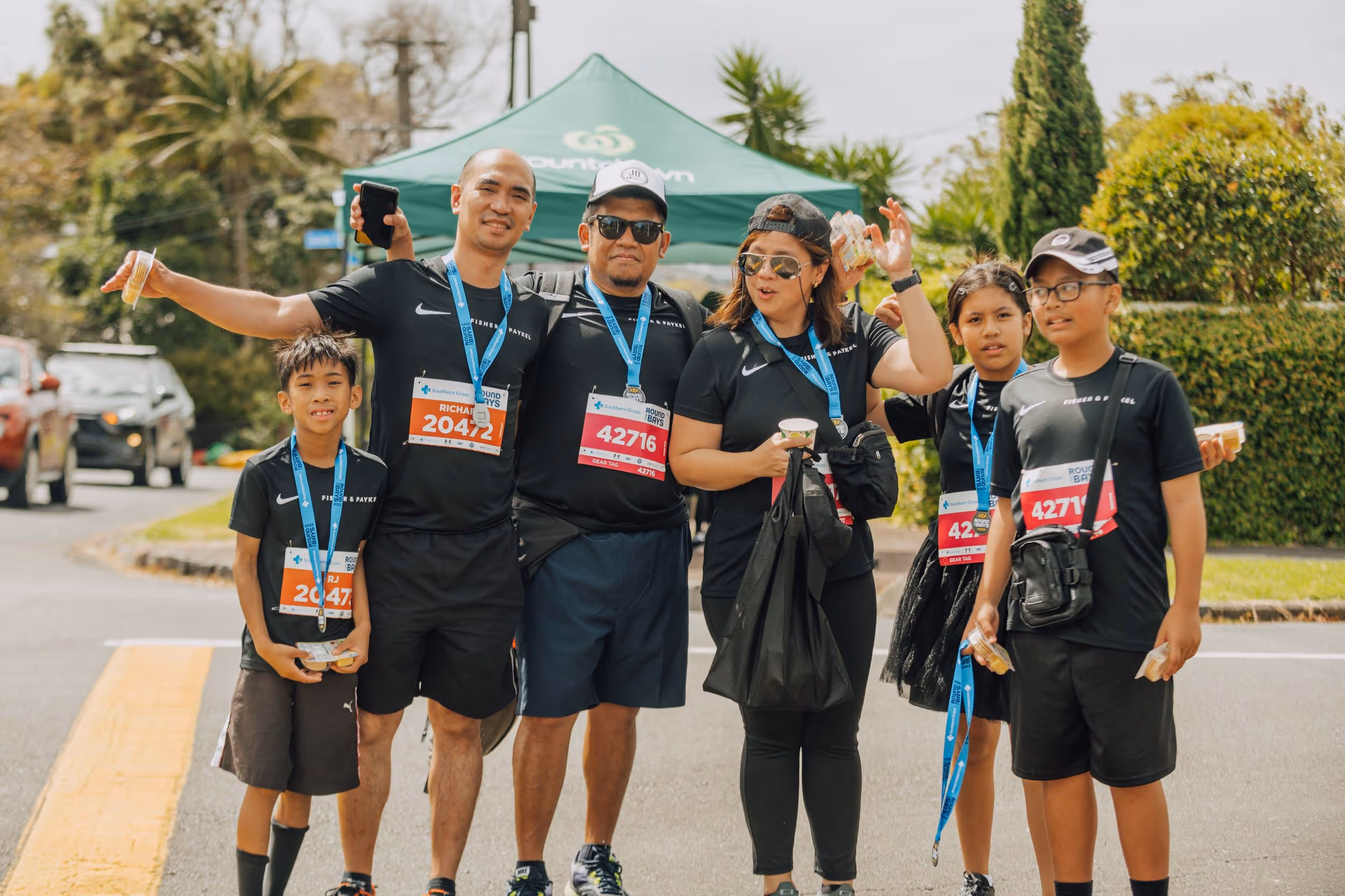 A group of six people, including adults and children, pose at an outdoor event. They all wear black shirts with race numbers and medals around their necks. Some of them hold water bottles and snacks. A large tent and trees are visible in the background.