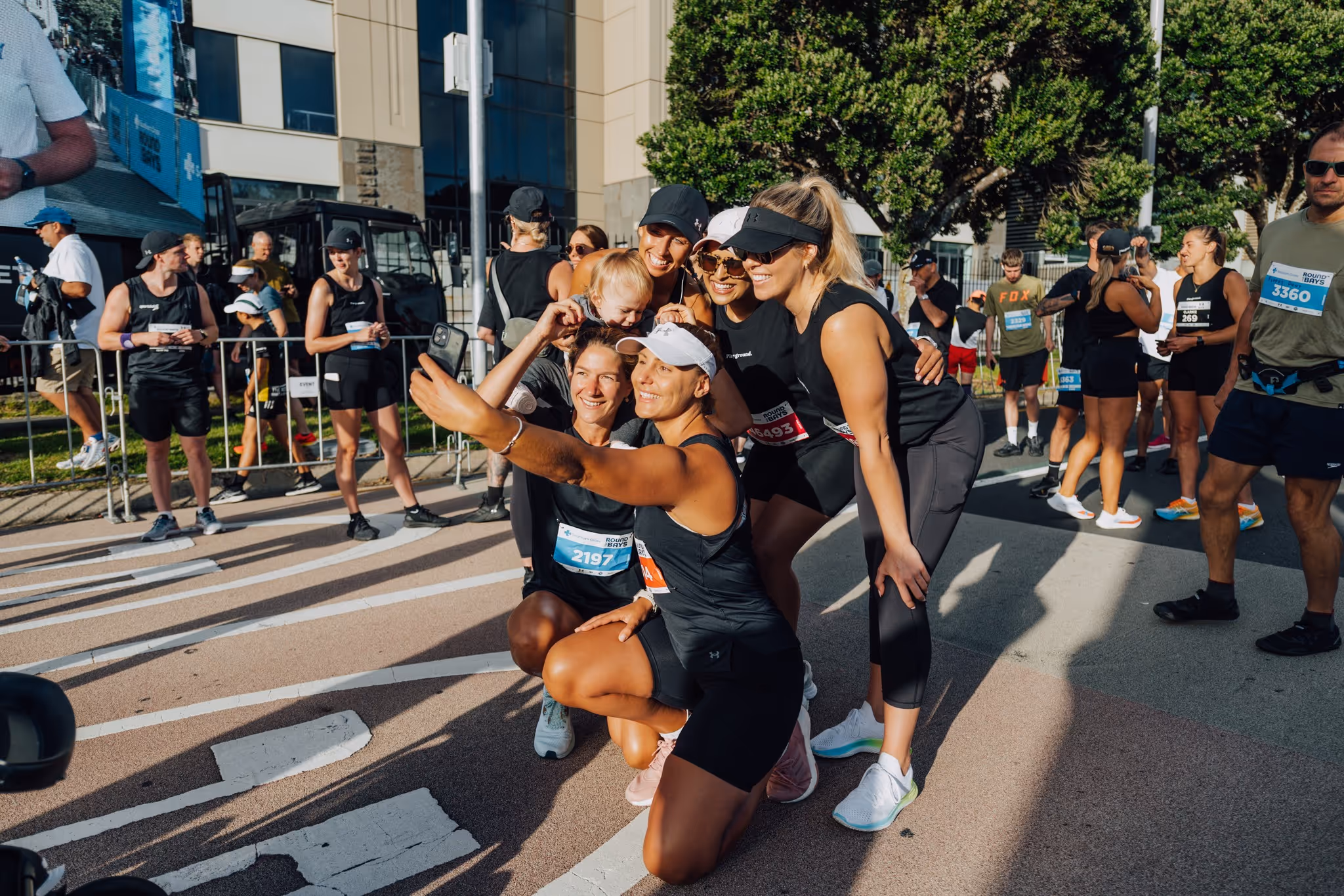 A group of women in athletic attire take a selfie during an outdoor event. They are surrounded by other participants, who are also dressed in activewear. The event appears to be in an urban area with modern buildings in the background.