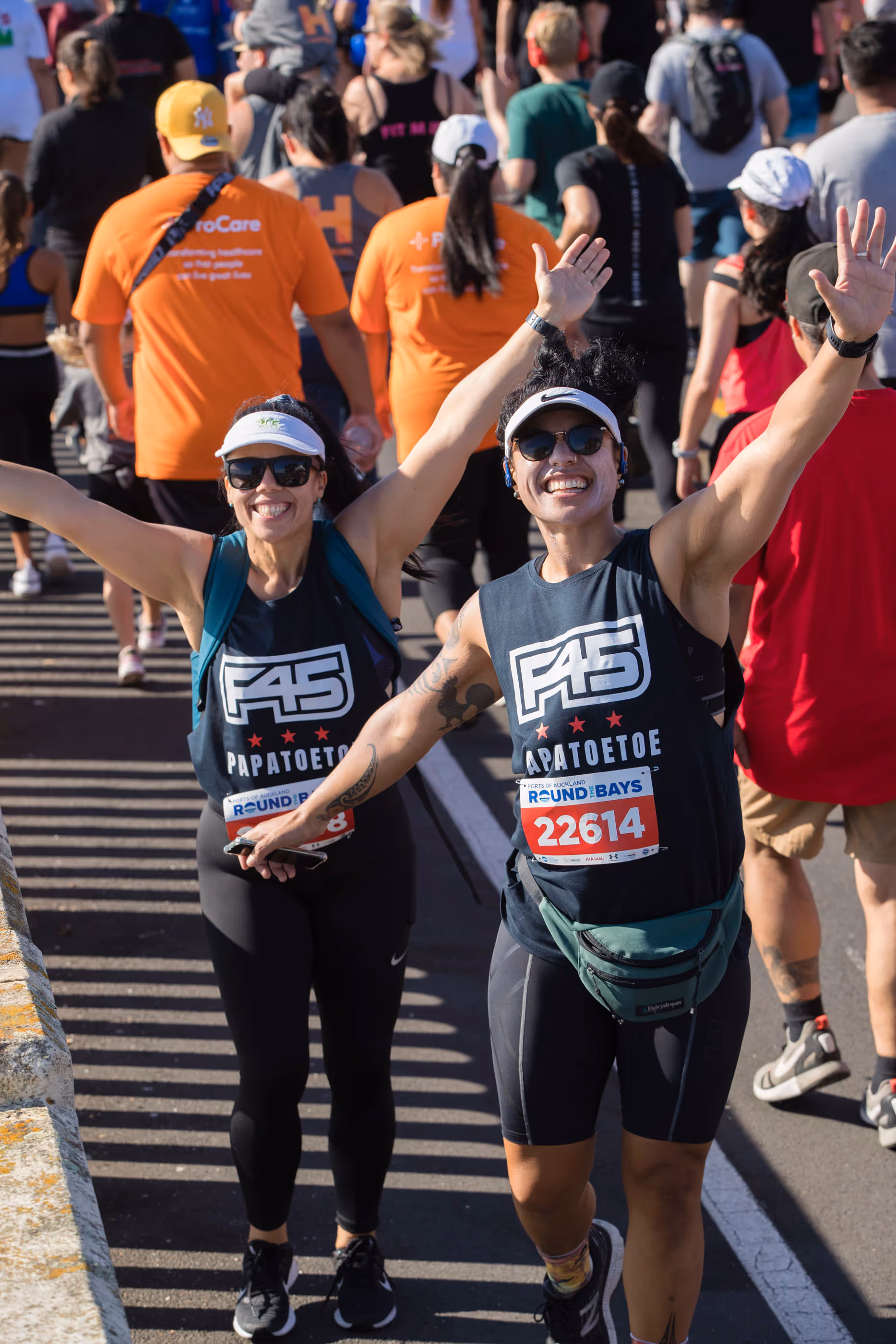 Two runners, a man, and woman, wearing athletic gear and sunglasses, smile and raise their arms in celebration as they near the finish line of a race. Other participants and spectators are visible in the background, creating a lively atmosphere.