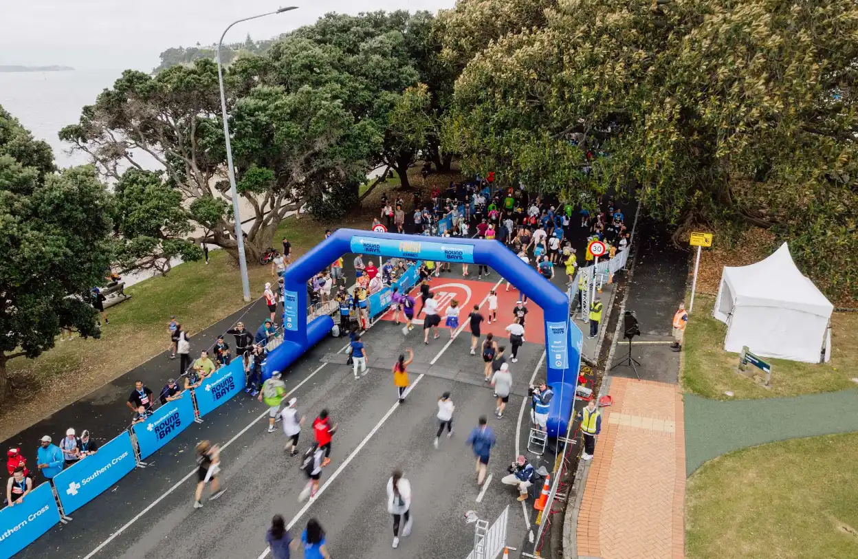 Aerial view of a marathon event with runners passing under a blue inflatable arch. The road is flanked by trees on one side and a grassy area with a white tent on the other. Spectators are lined along the sides of the road. The sky appears overcast.
