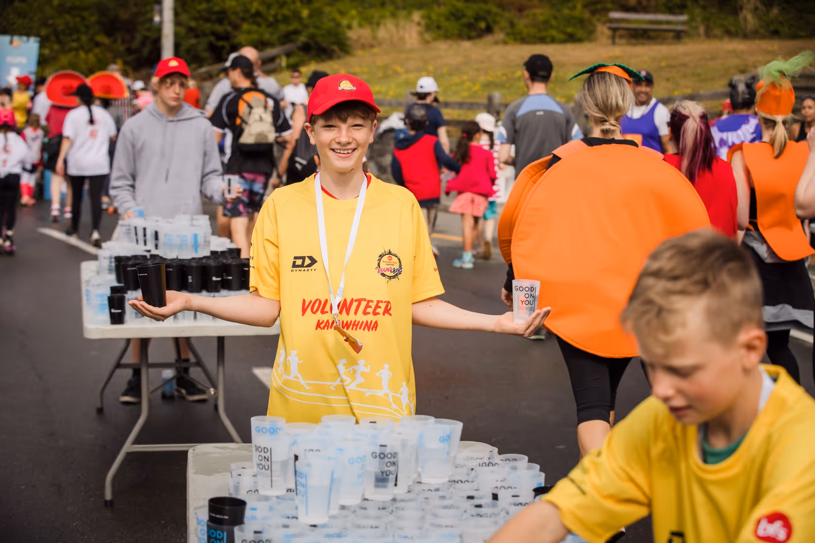 Volunteers at a race event. A young boy in a red hat and yellow shirt labeled "race official" holds cups of water. Another child, also in a yellow shirt, handles water cups on a table. Participants in race attire are visible in the background.