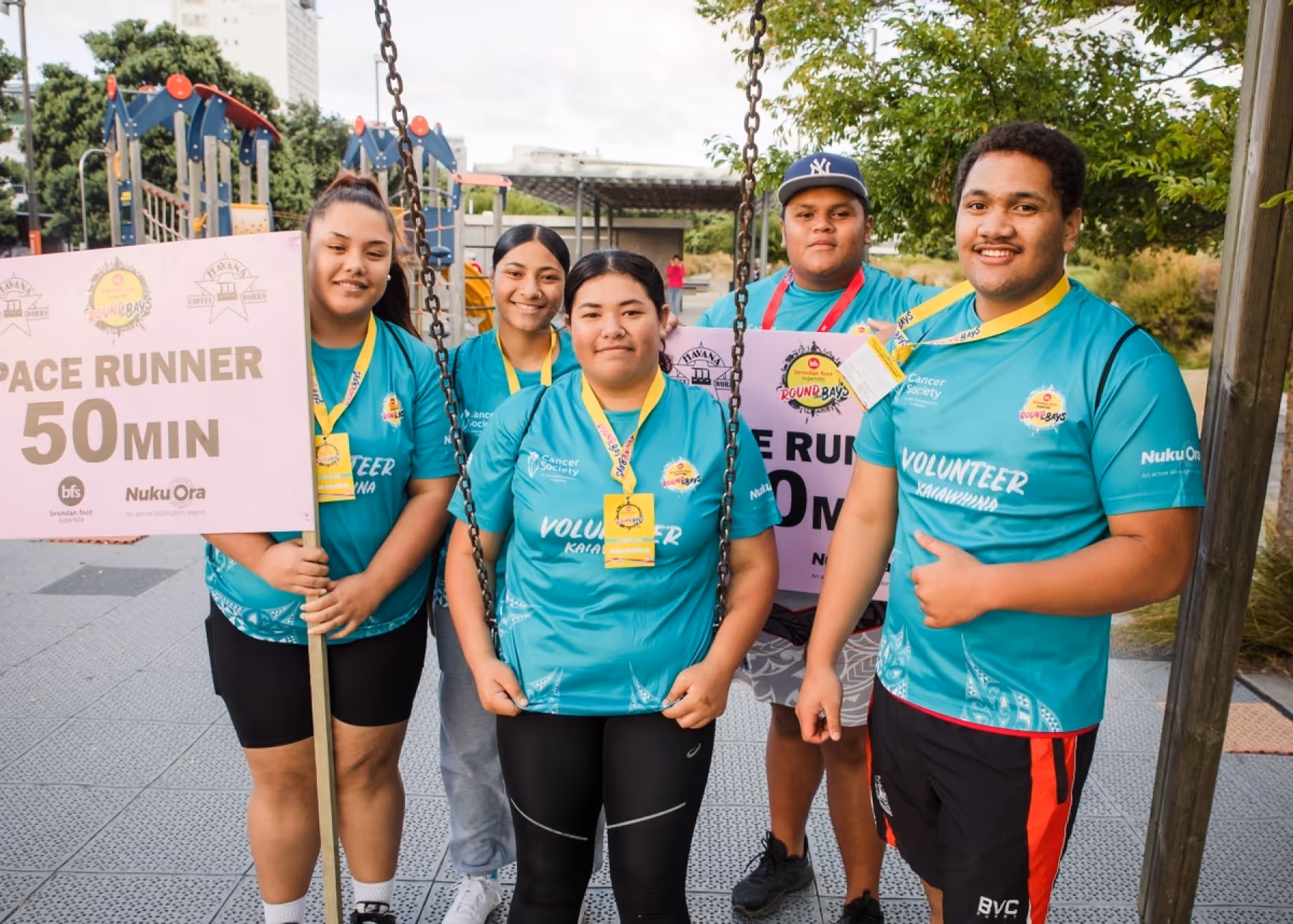 A group of five people wearing teal shirts with lanyards around their necks pose together after volunteering at a running challenge. They are standing outdoors, and a sign in the background reads, "50 MIN." The atmosphere is cheerful and celebratory.