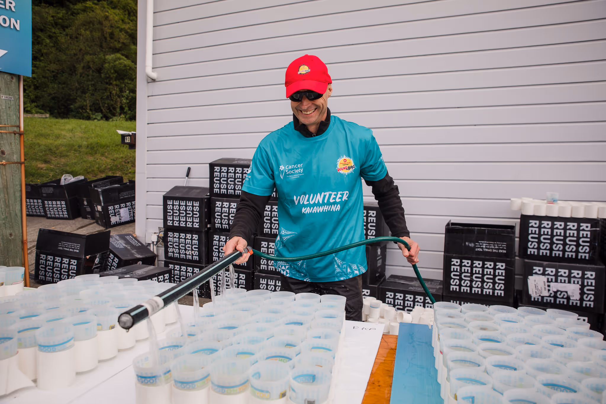 A person in a red cap and teal shirt is topping up cups of water for runners and walkers at an event