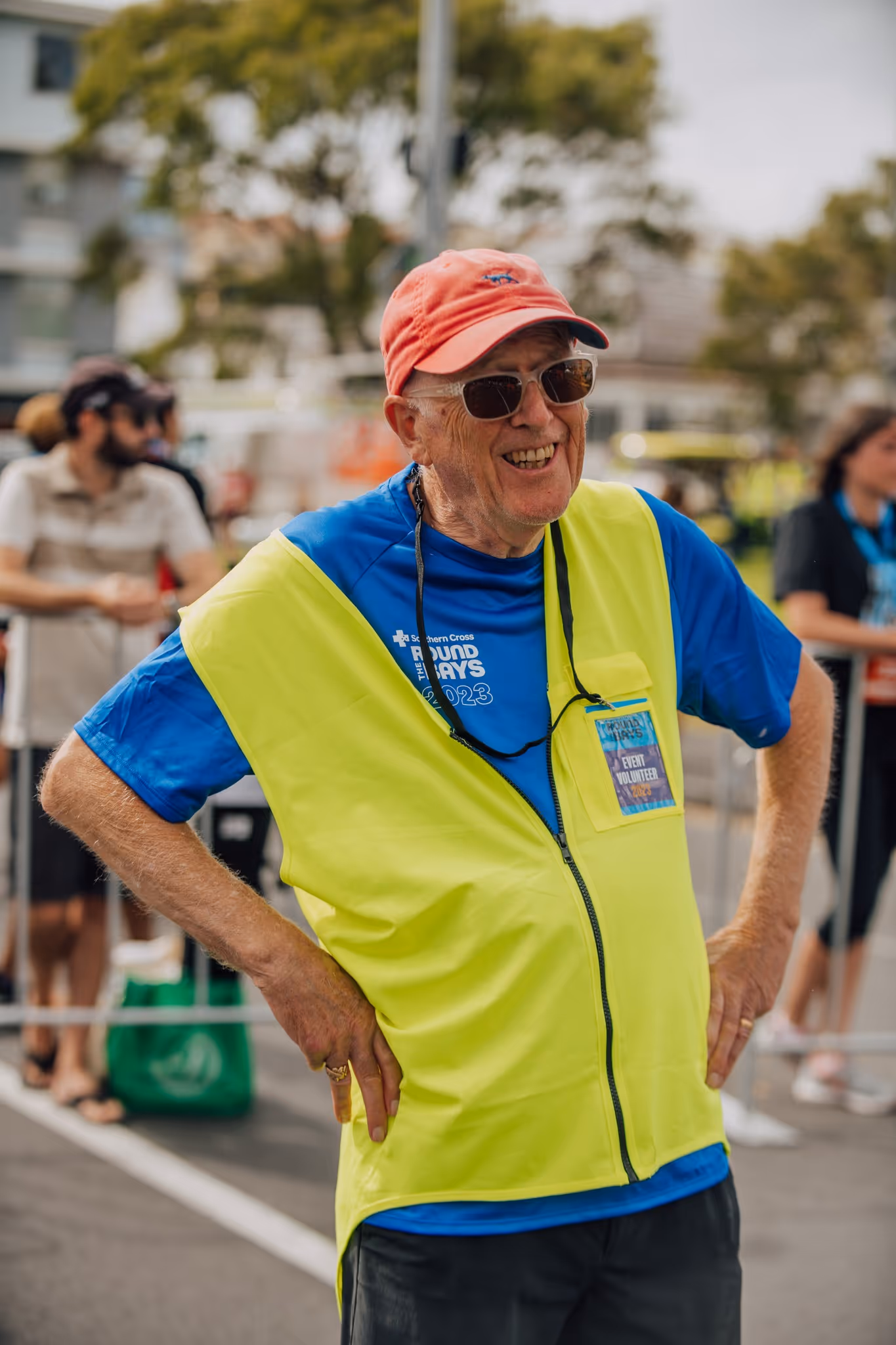 A man wearing a yellow safety vest, blue shirt, red cap, and sunglasses stands with his hands on his hips. He has a whistle around his neck and a badge clipped to his vest. Several people are in the background, some of whom are behind a barrier.