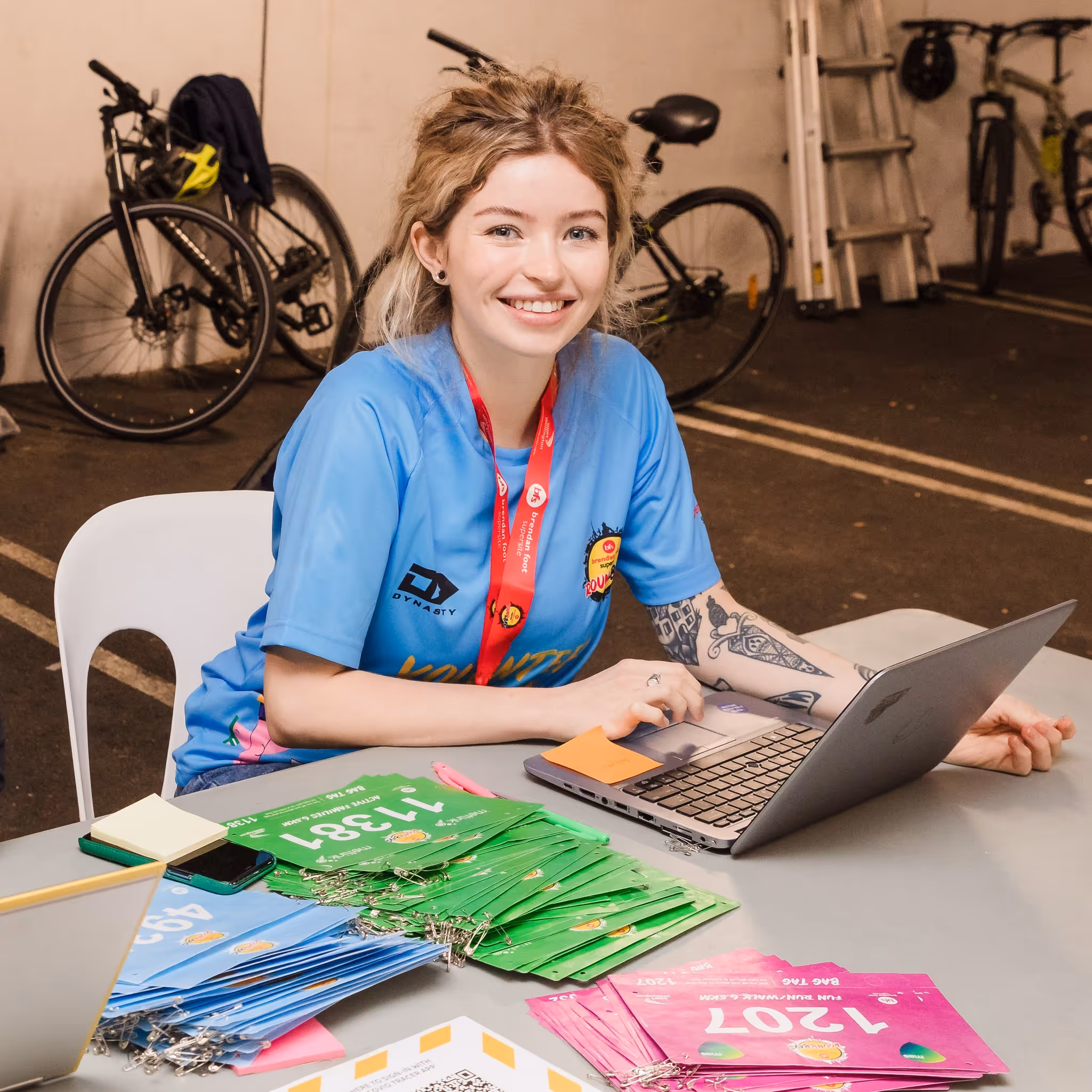 A smiling person in a blue shirt sits at a table with a laptop and colorful leaflets, including green and pink ones. Behind them, there are parked bicycles. The setting appears to be indoors, possibly a registration area for an event.