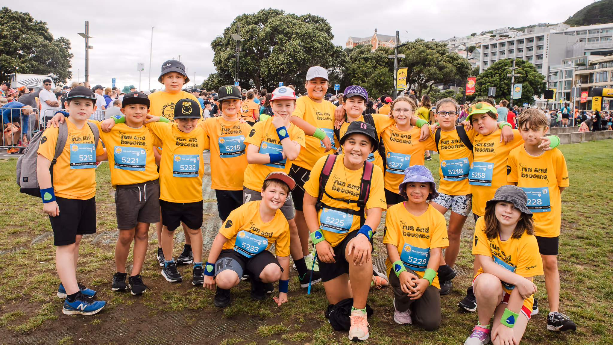 A group of children wearing yellow shirts and race bibs gather for a group photo outdoors. They appear to be participating in a charitable or sports event. The background features trees and buildings, and some participants are holding balloons.