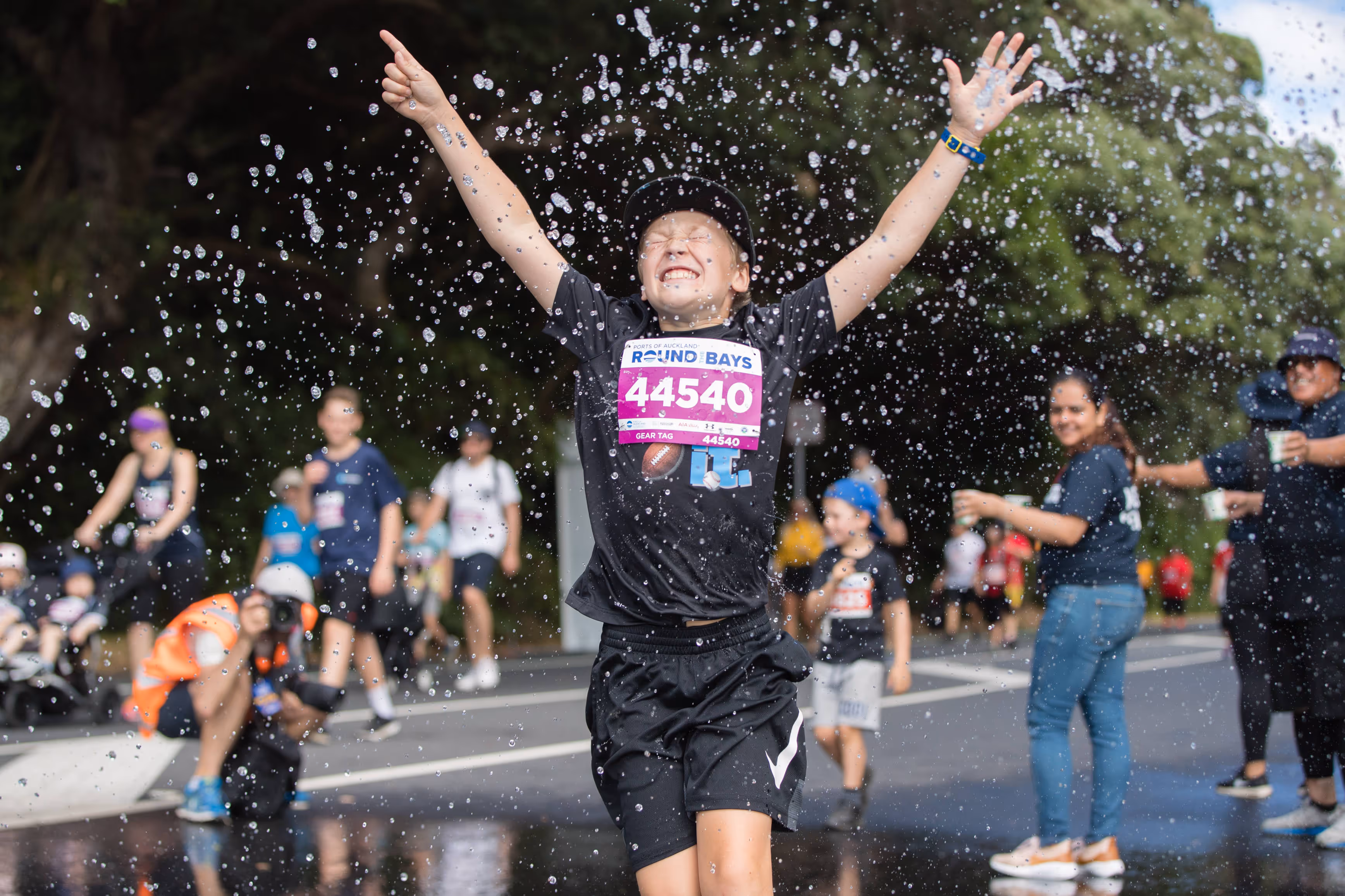 A joyful runner wearing a "STRONG MIND STRONG BODY" T-shirt crosses the finish line with arms raised in celebration. Other participants and spectators cheer in the background on a sunny day with trees lining the street.