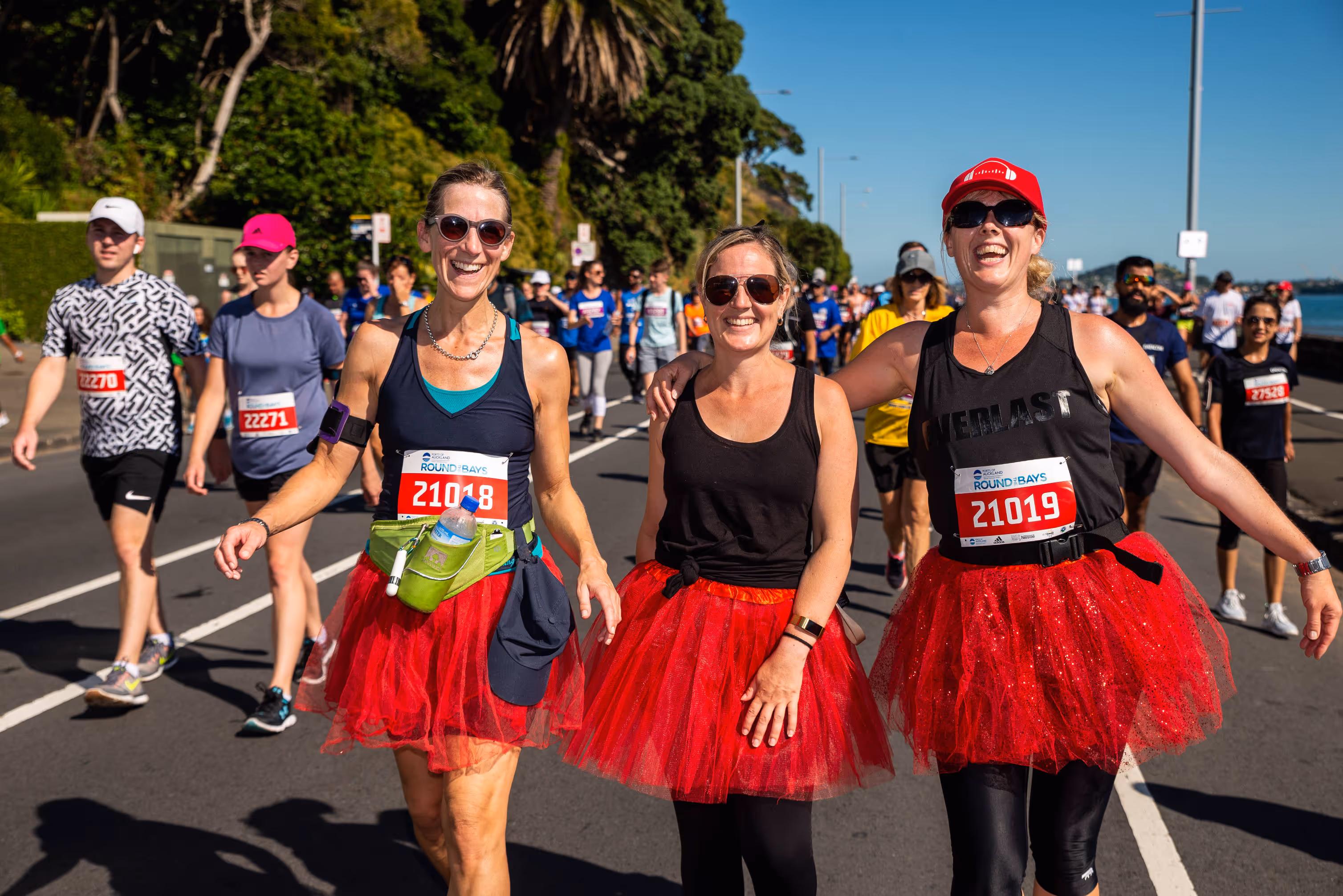 A group of runners participates in a race. Three women in the foreground wear red tutus and black tops, smiling and holding hands as they run. People run on the road in the background amidst a sunny, scenic environment with greenery and a clear sky.