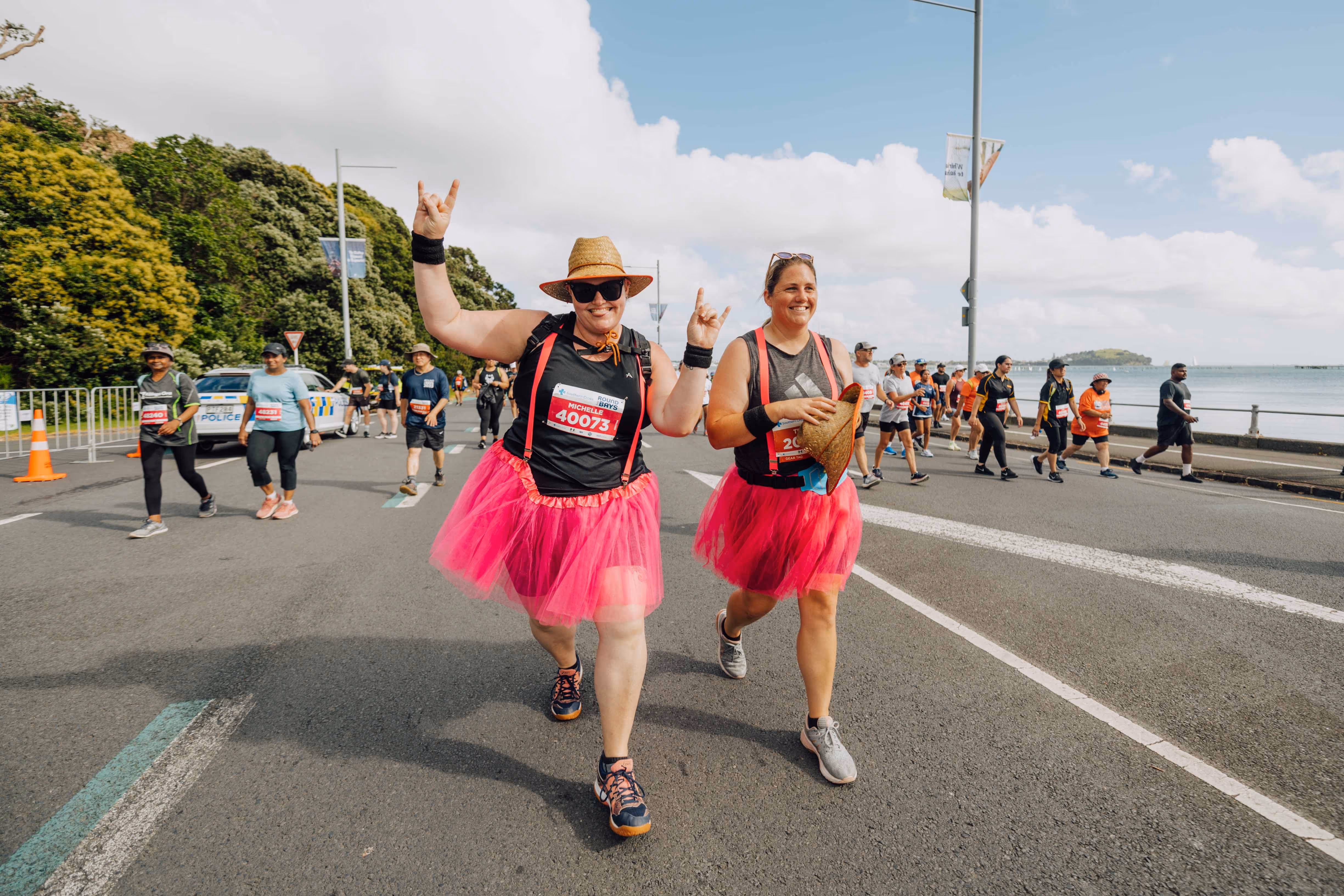 Two people in pink tutus run on a road during a race or fun run event. Both are smiling and have their arms raised in a celebratory manner. Other participants and spectators can be seen in the background. It's a sunny day with a few clouds in the sky.