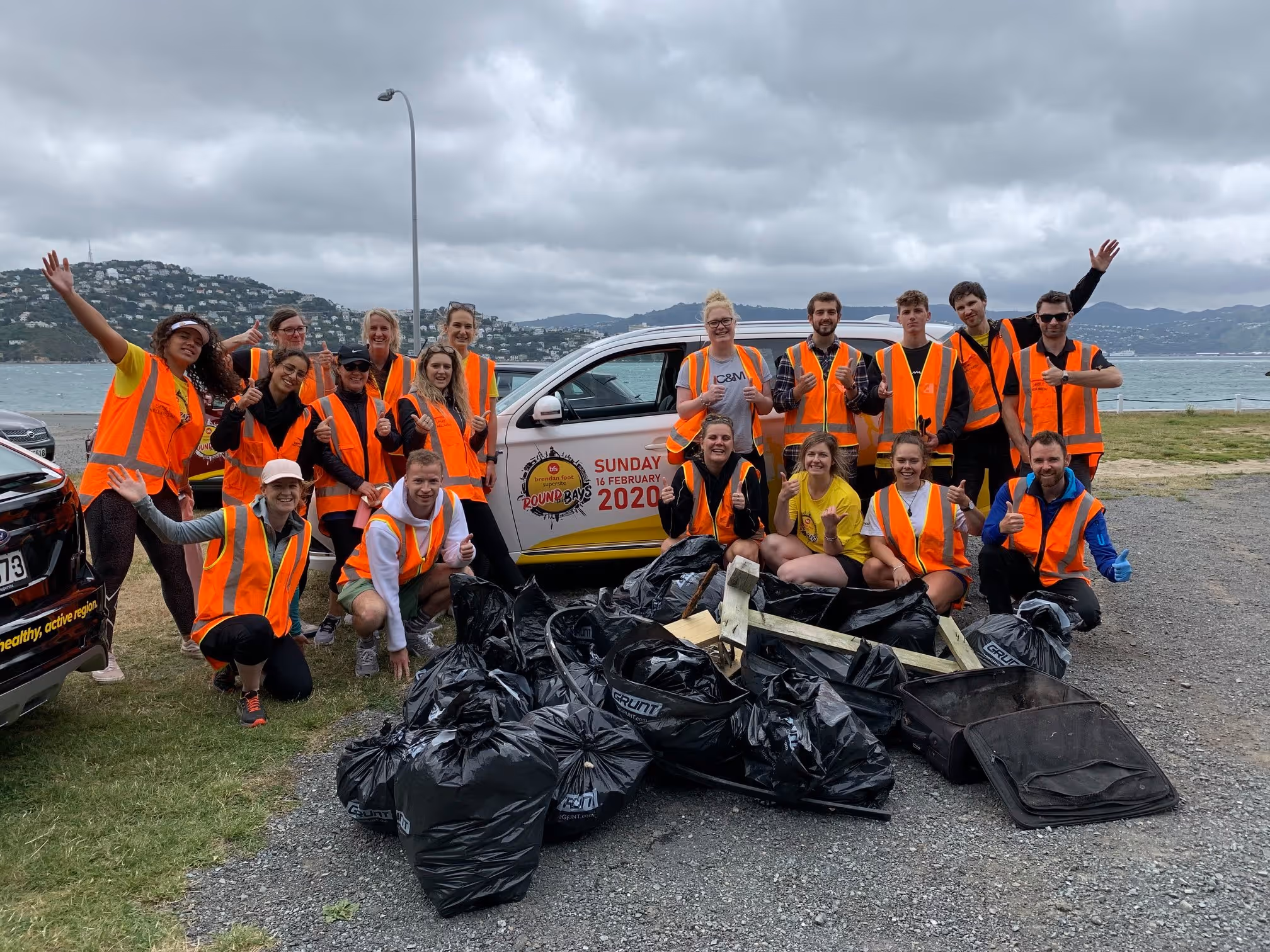 A group of people in orange safety vests pose for a photo outdoors by the water, with a cloudy sky overhead. They are standing and kneeling behind a collection of filled garbage bags and litter they have gathered, with a patrol vehicle visible in the background.