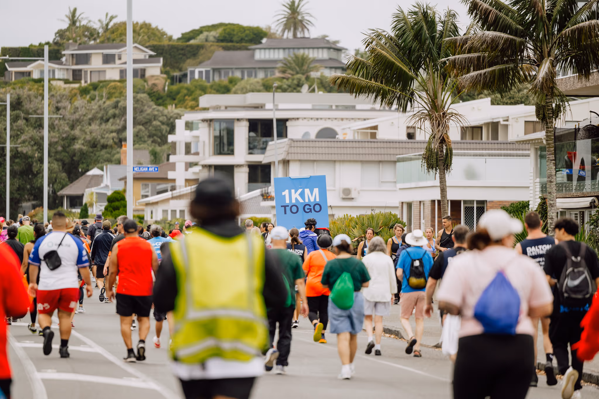 Participants dressed in athletic gear are walking or running on a road lined with buildings and trees during an outdoor event, with some wearing high-visibility vests and bibs. It's a cloudy day, and the road is surrounded by greenery and parked cars.