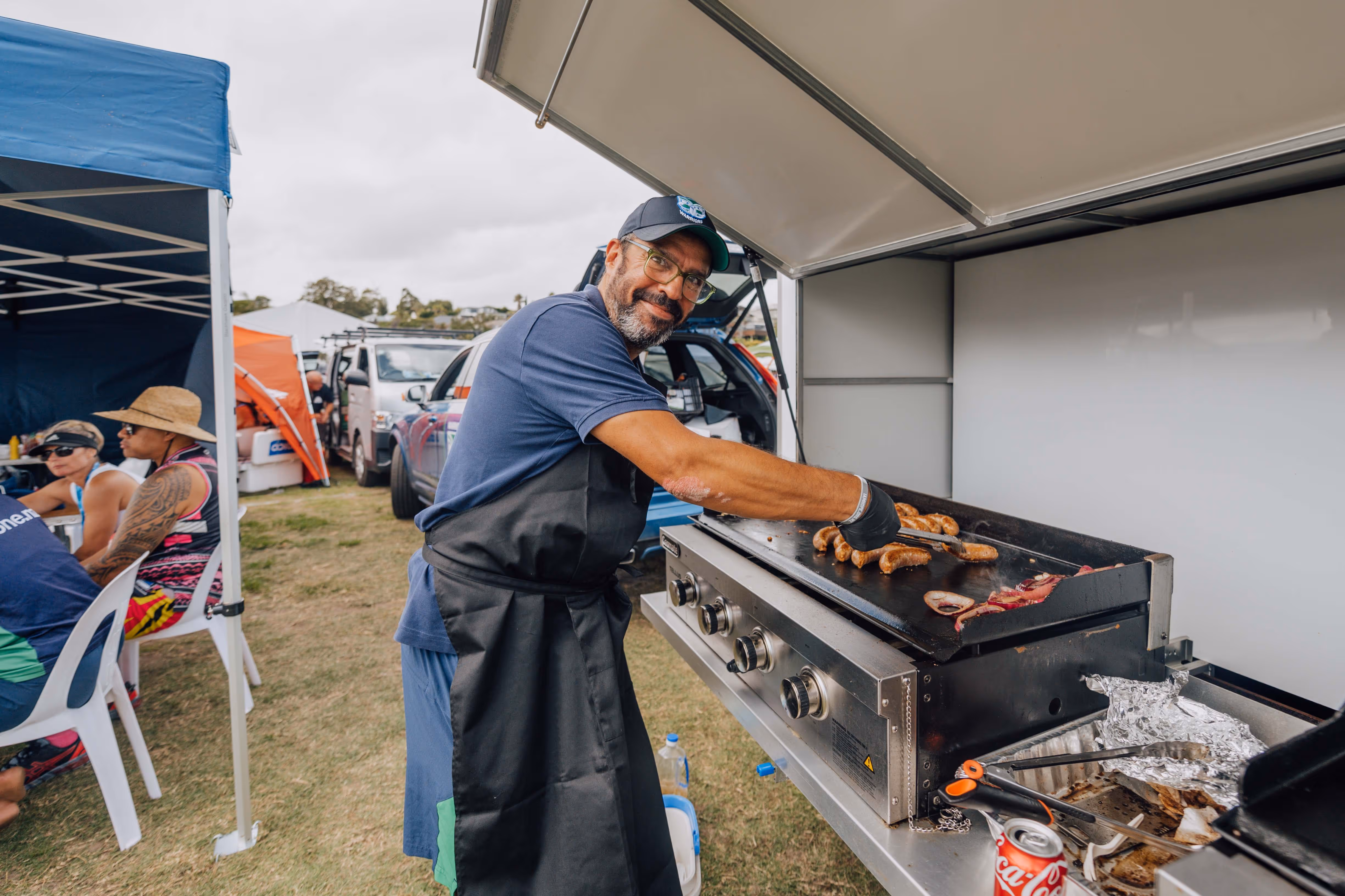A person in a blue shirt and apron cooks meat on a large outdoor griddle at a community event. Several people are sitting and eating under a nearby tent, with vehicles and other tents in the background.