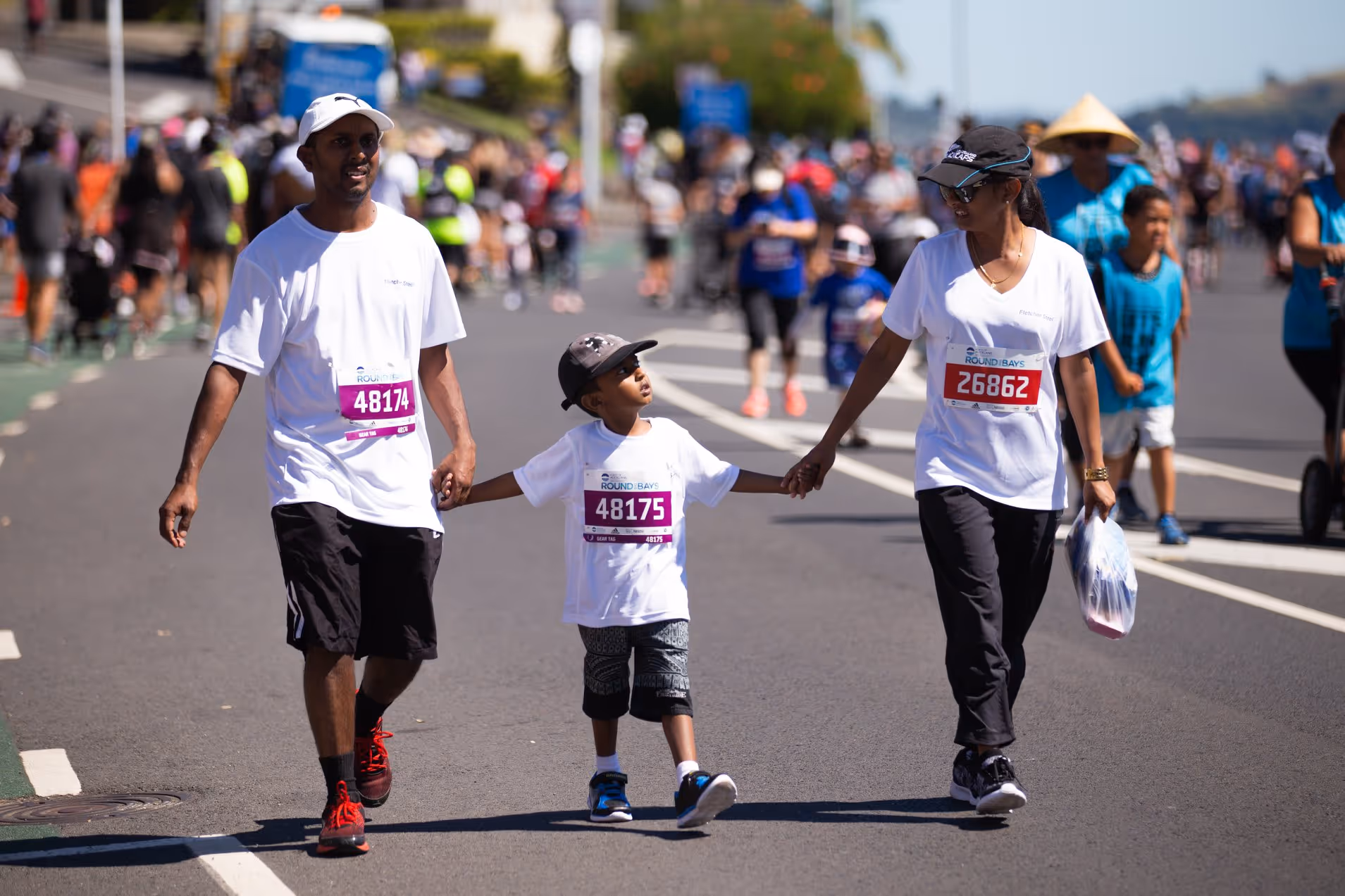 A smiling family of three, wearing matching white shirts, holds hands while walking on a street during a race event. Many other participants and spectators can be seen in the background under a sunny sky.