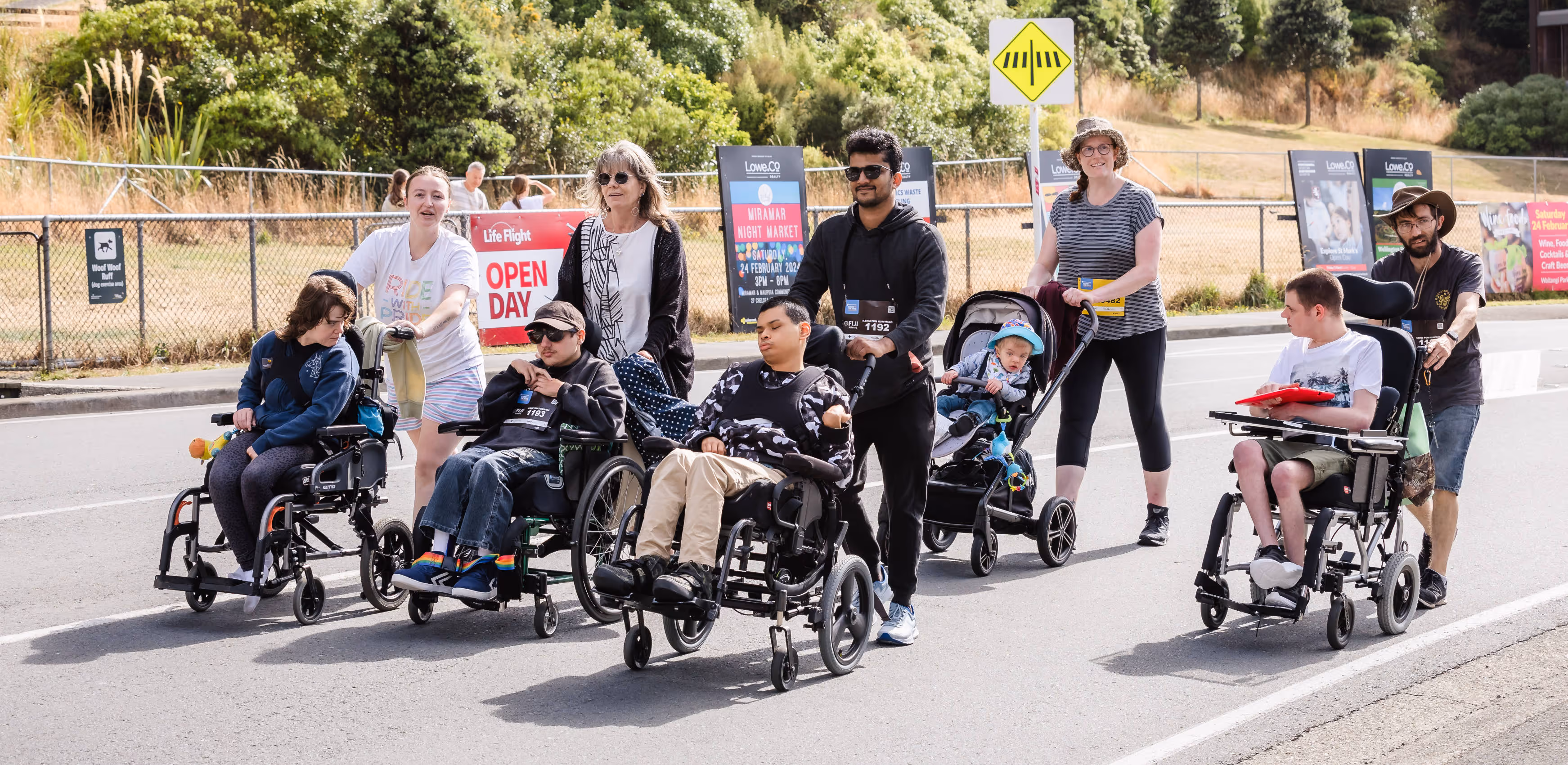 A group of people, including children and adults, are on a walk. Some of them are using wheelchairs. They are outdoors on a paved pathway with greenery and trees in the background. Everyone appears to be engaged and enjoying the walk.