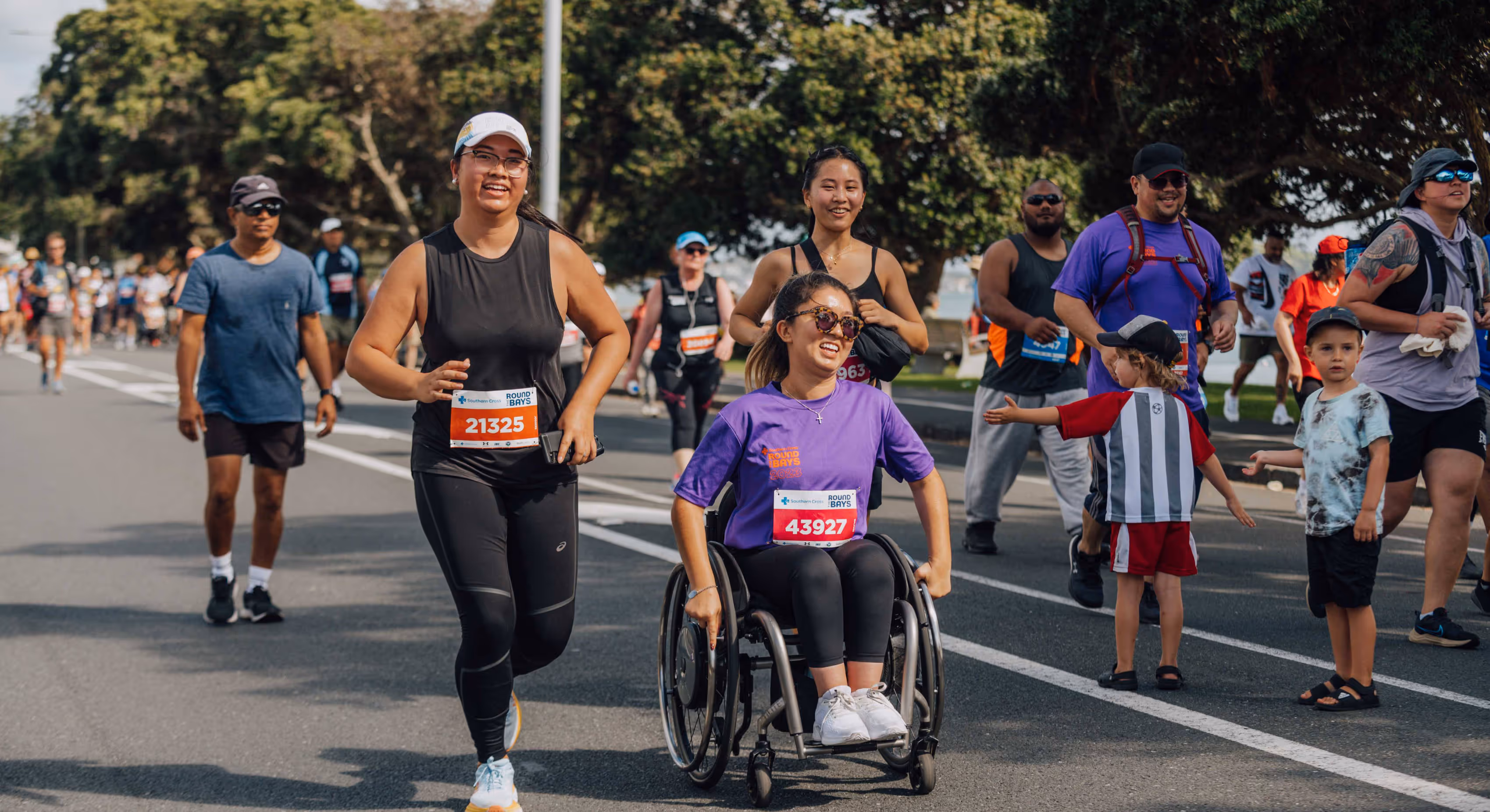 A diverse group of participants in athletic wear are running and wheeling down a street in a race. One person in a wheelchair wearing a purple shirt is being pushed by another participant. Spectators line the side of the road cheering them on.