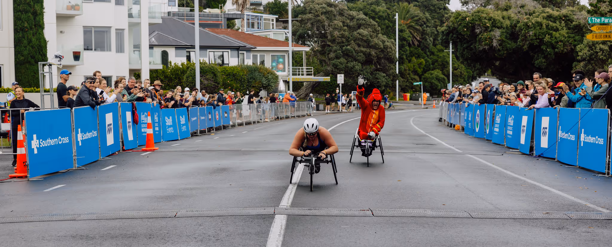 Two athletes compete in a wheelchair race on a city street lined with blue barricades and spectators. The athlete in the foreground is pushing with intense effort, while the other follows closely behind. Residential buildings and trees are visible in the background.